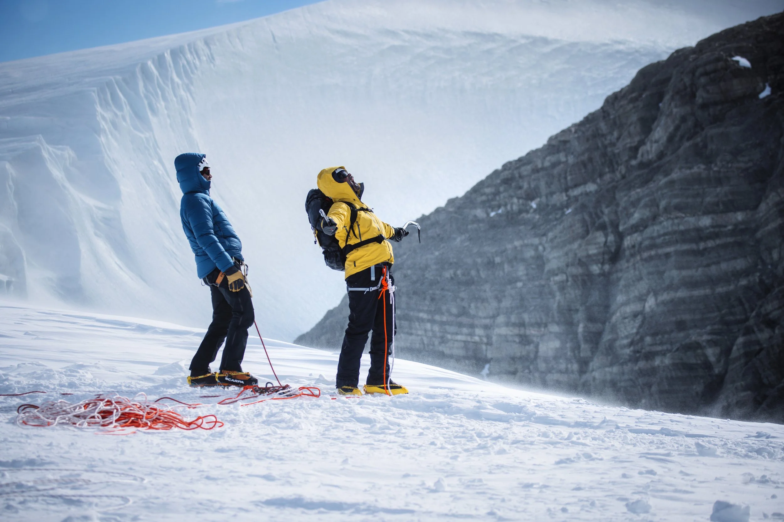 Will Smith & Friend dressed in winter gear standing on snowy terrain with a large ice wall and rocky cliffs in the background.