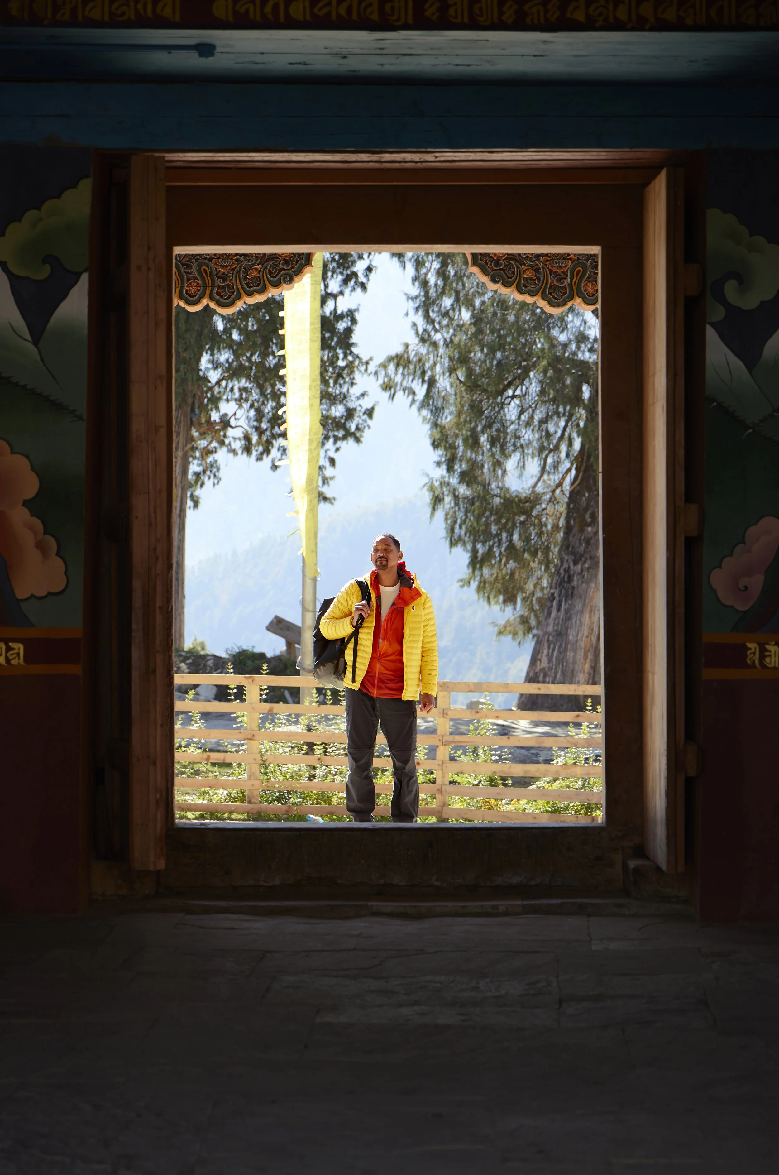 Will Smith wearing a yellow jacket and red shirt standing outdoors on a wooden platform, looking up, with trees and mountains in the background, seen through a framed doorway.