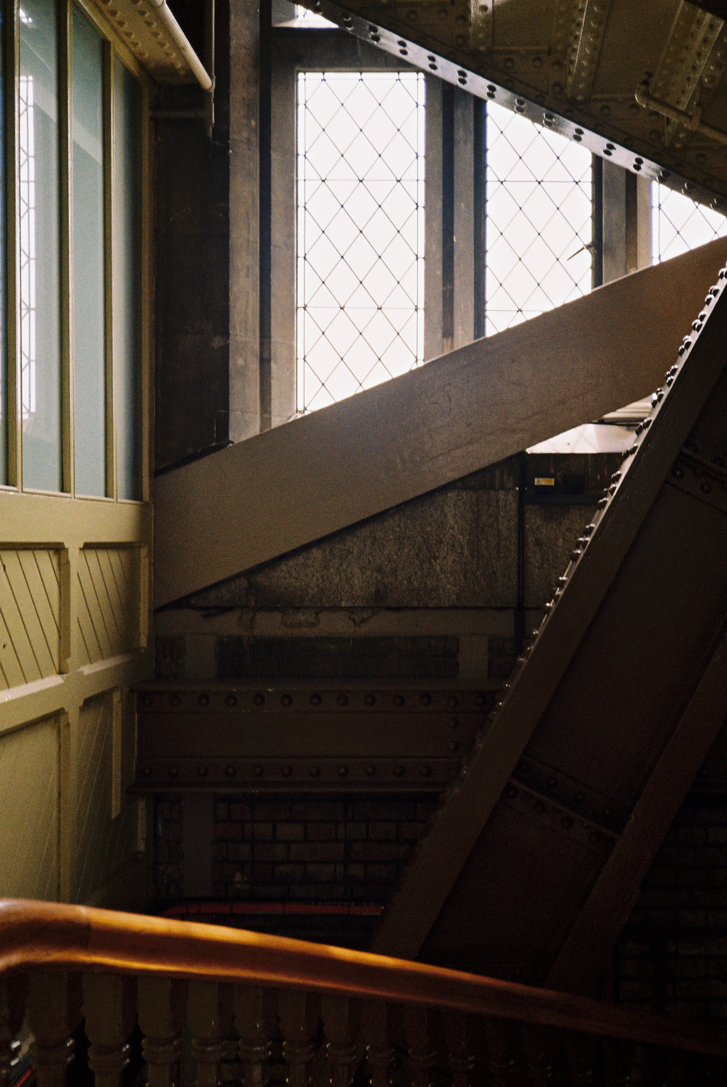 View of an indoor staircase with wooden railings and decorative metalwork, leading up to a large window with grid-like panes letting in natural light.  Photographed by Julia Tran in London, United Kingdom. Photographed on 35mm film. 