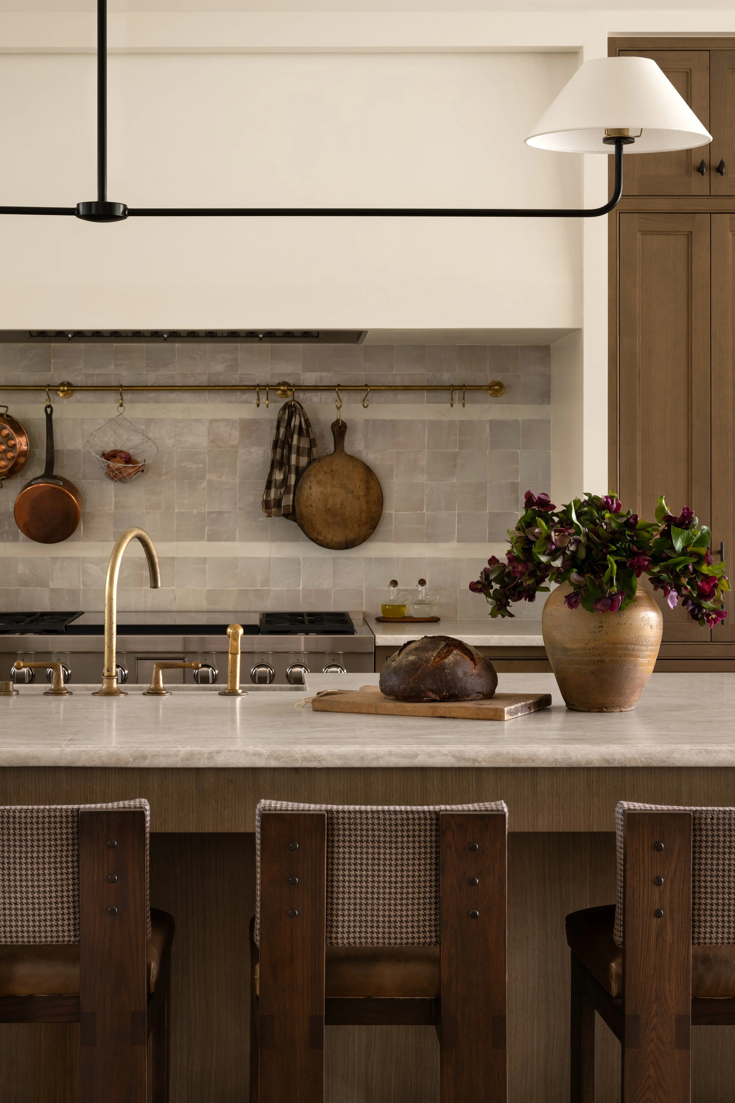 In a space designed by Studio McGee, Modern kitchen with beige marble countertop, hanging copper pots, a round wooden cutting board, a large rustic loaf of bread, a beige urn with purple flowers, wooden bar stools, and cabinetry.