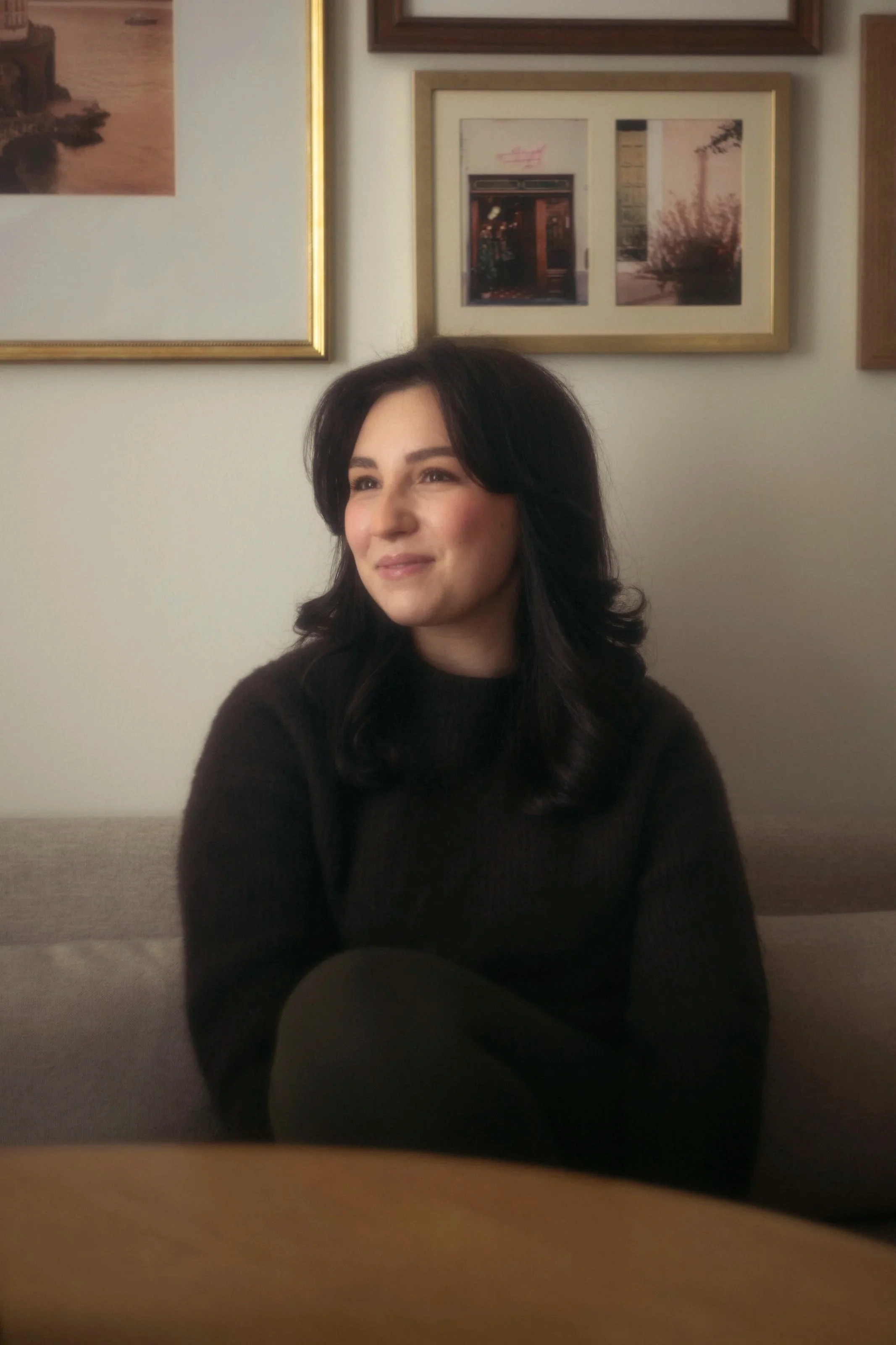 A woman with dark hair is sitting in a room with a beige wall. Behind her, there are framed pictures hanging on the wall.
