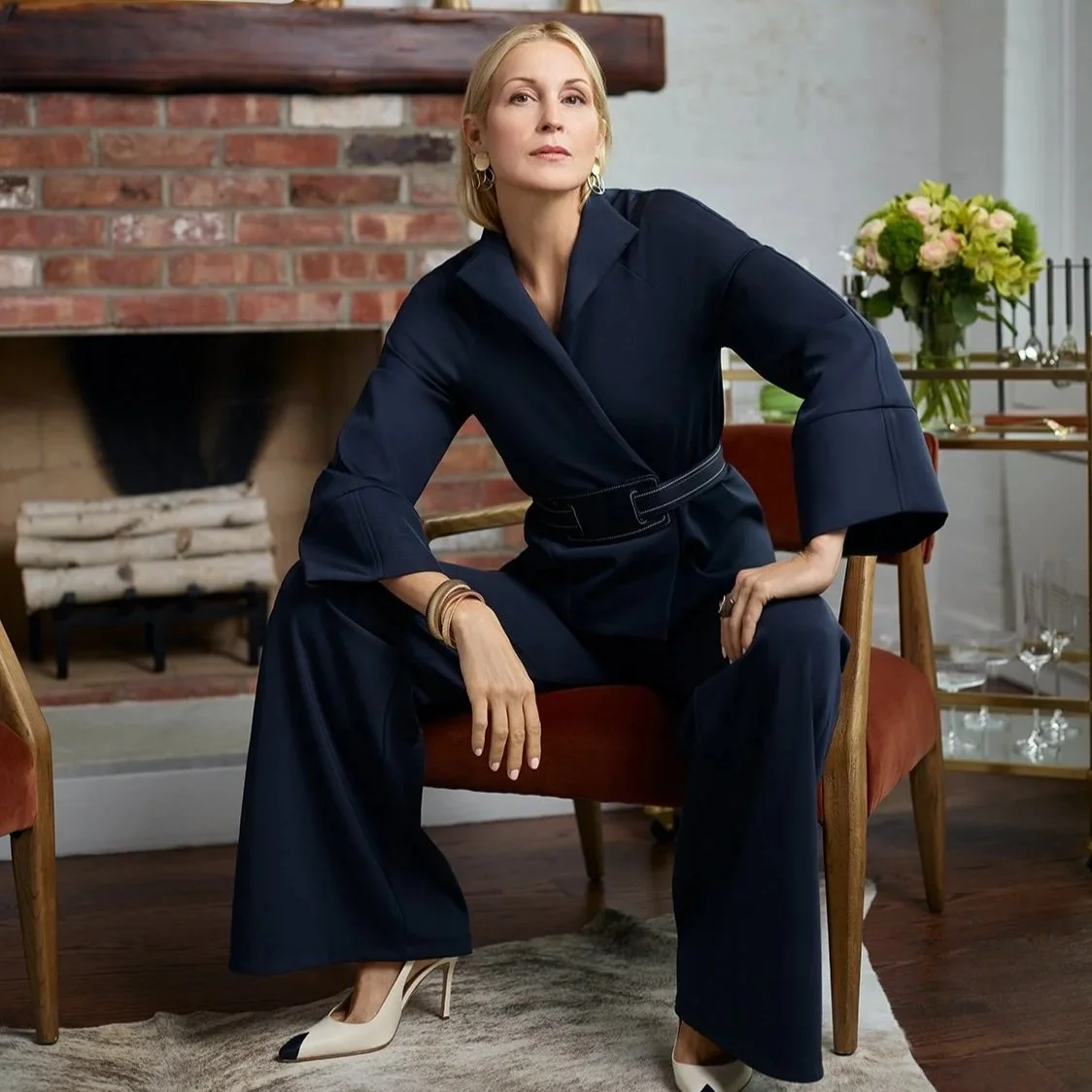 Kelly Rutherford wearing a dark navy jacket and wide-leg pants, sitting on a wooden chair in a room with a brick fireplace, a vase of flowers, and glassware in the background for SITA.