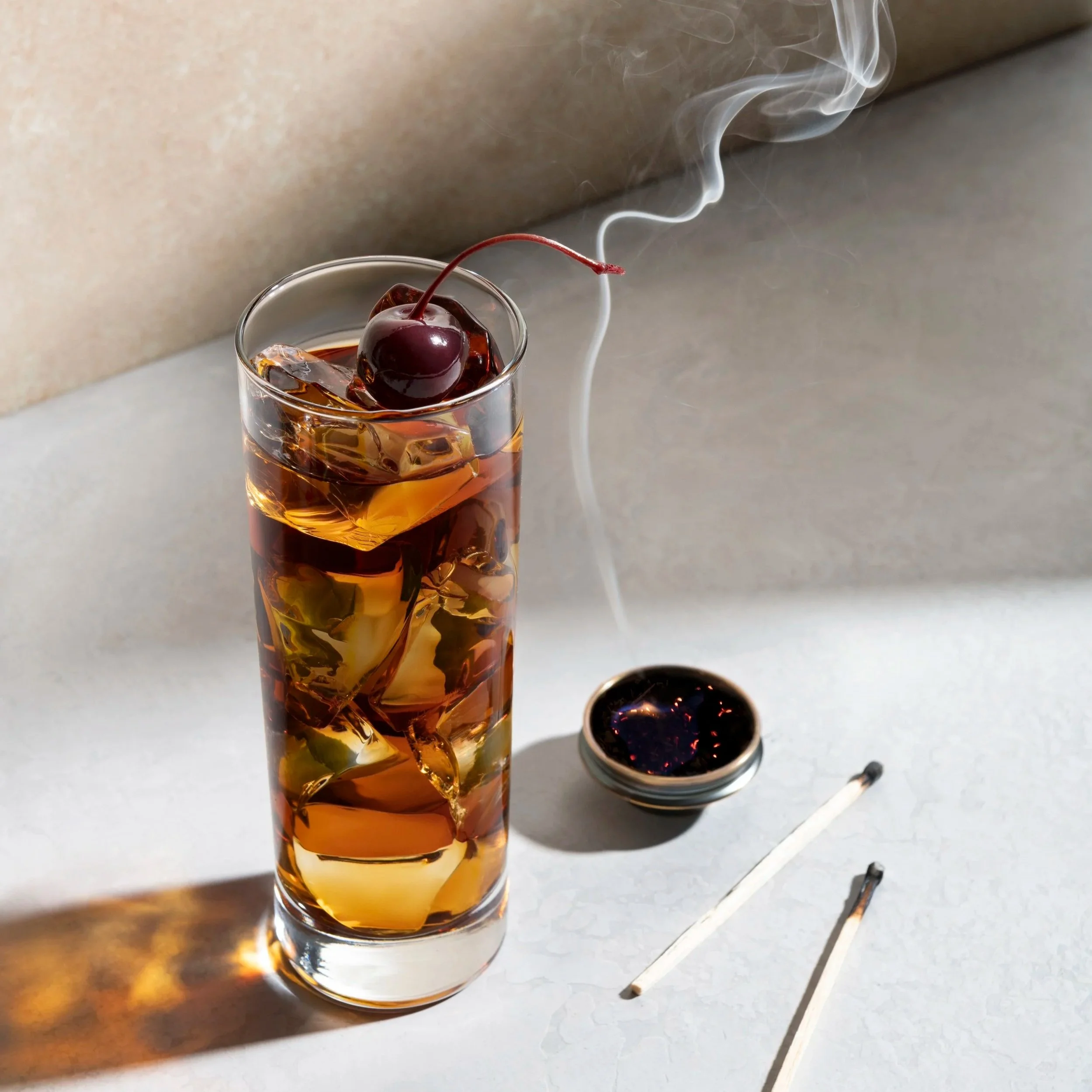 A glass of whiskey with ice cubes and a maraschino cherry inside, placed on a white surface with a shadow cast on it. A small bowl containing dark liquid and some metal utensils are also visible on the surface.