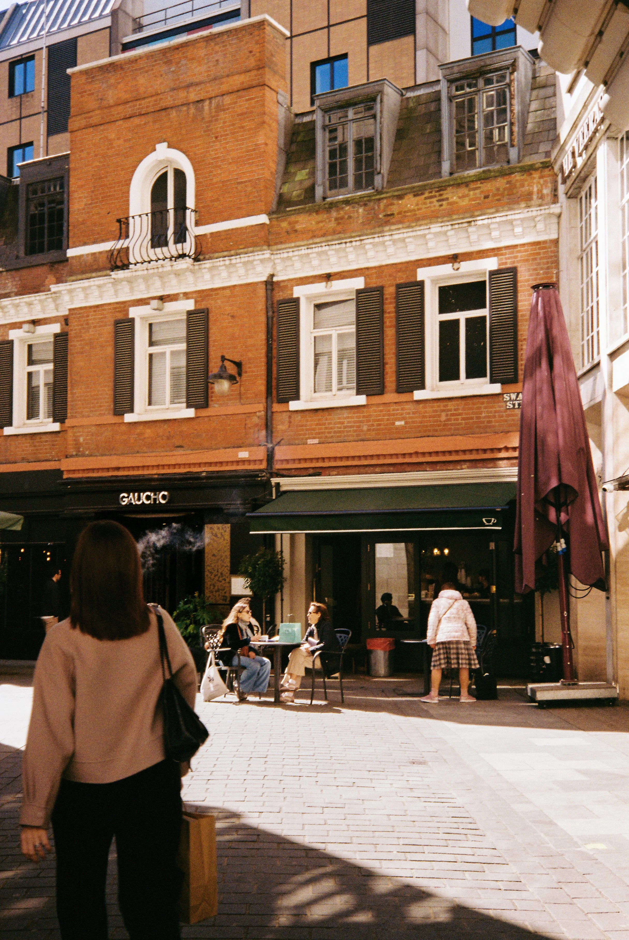 People sitting and walking outside a cafe on a city street, with brick and stone buildings and windows with shutters in the background.  Photographed by Julia Tran in London, United Kingdom. Photographed on 35mm film. 