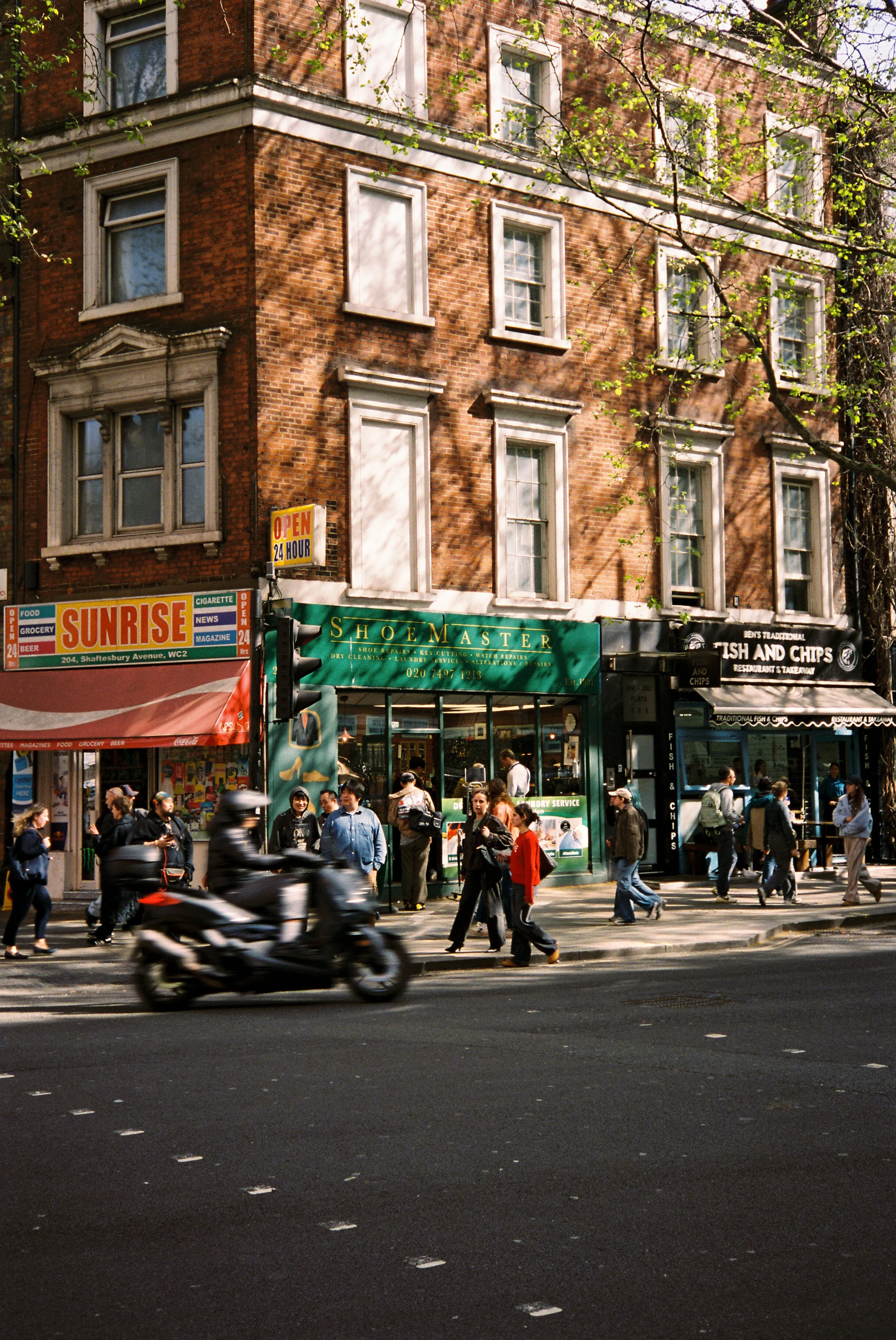 Busy city street with pedestrians crossing and riding bikes, storefronts with signs including Sunrise, Shoemaster, and Fish and Chips, and a brick building with multiple windows.  Photographed by Julia Tran in London, United Kingdom.