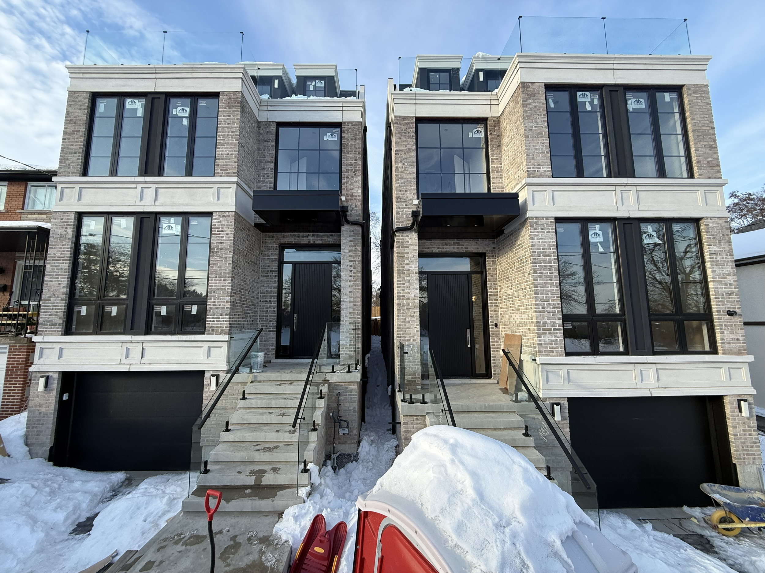 Two modern multi-story brick townhouses under construction with snow on the ground, featuring large black-framed windows, black doors, and exterior stairs with black railings.