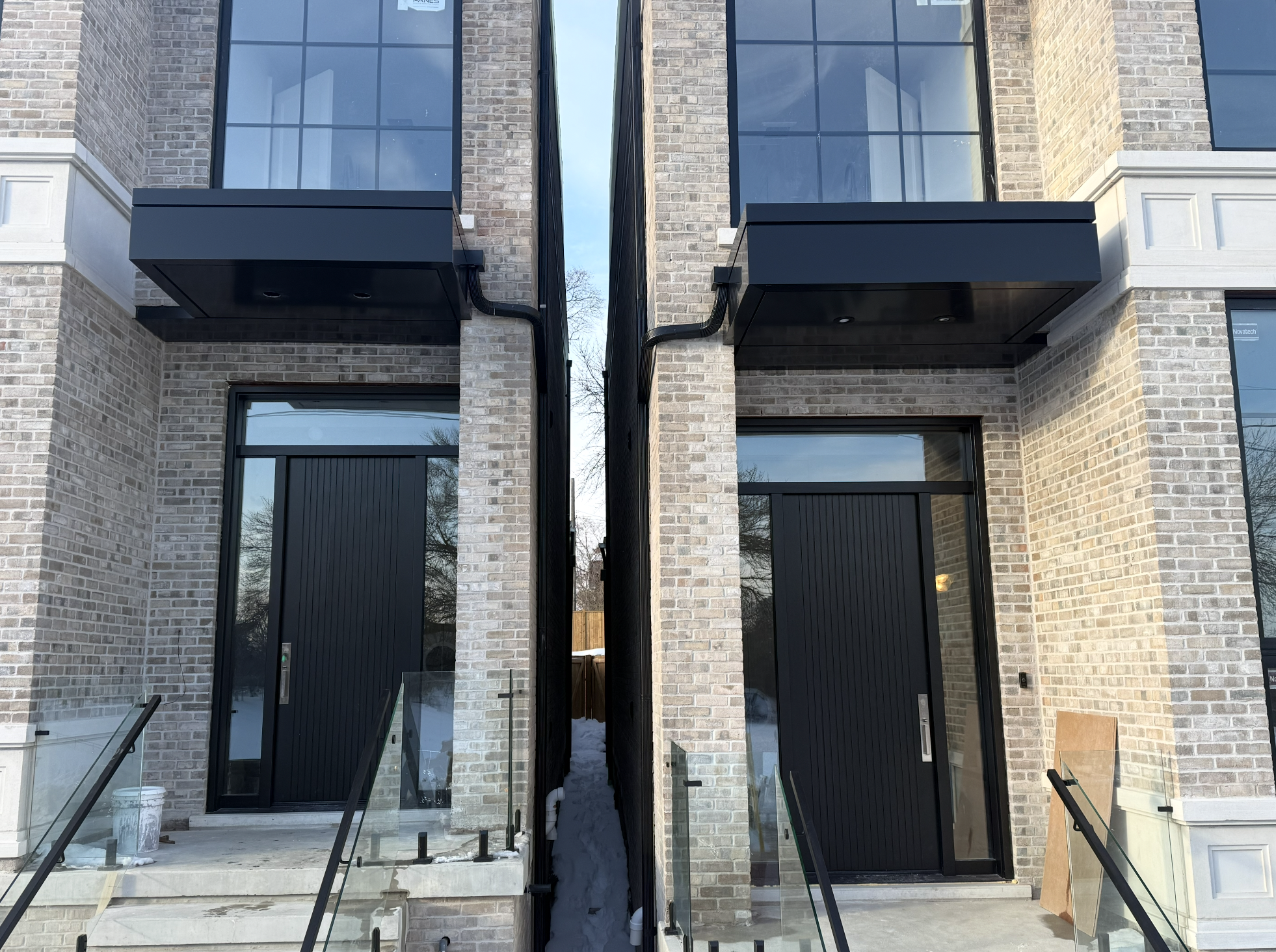 Two modern brick townhouses with glass front doors and black accents, separated by a narrow alley with snow, black awnings above doors, and glass railings on stairs.
