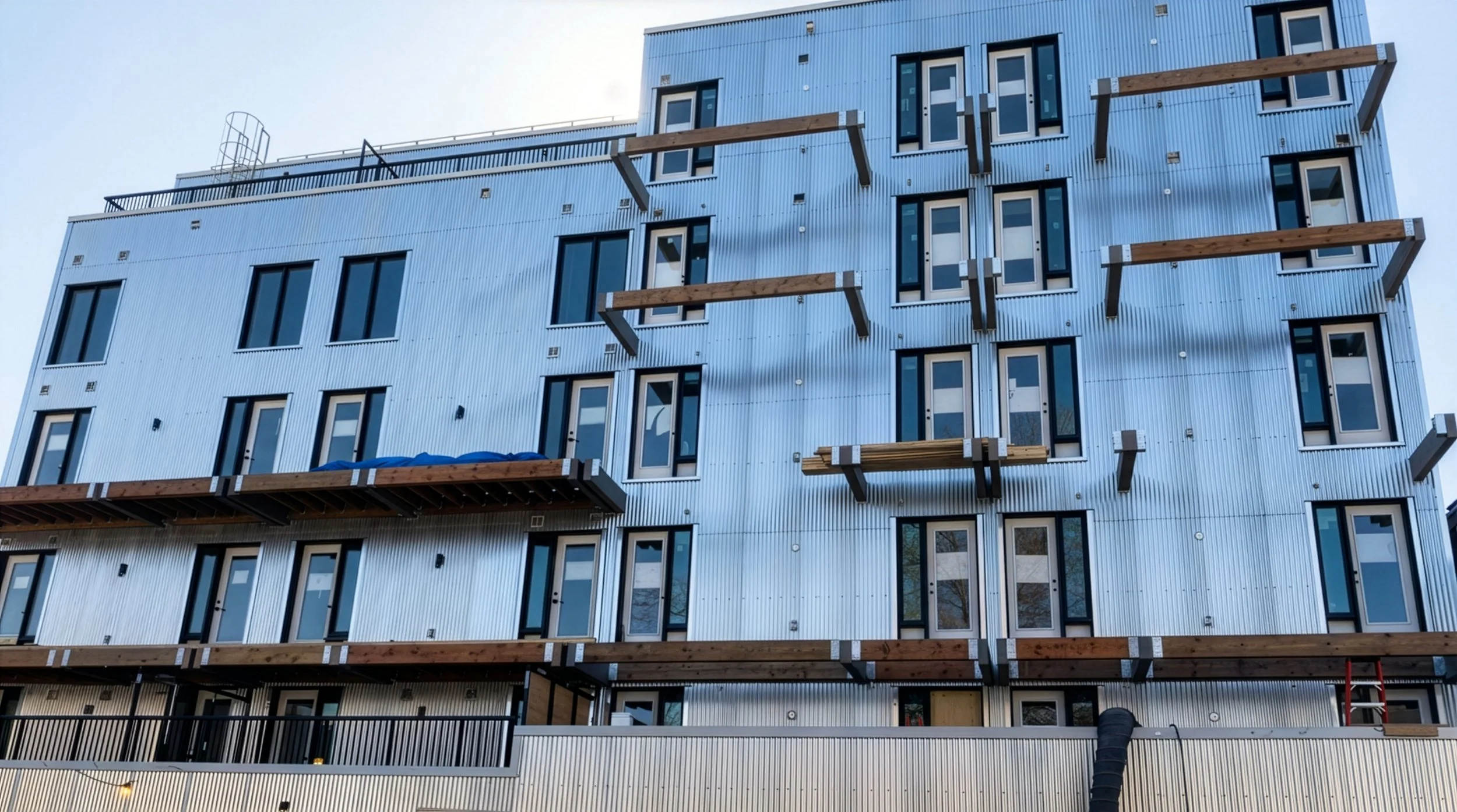 Under construction modern multi-story residential building with metal siding, multiple windows, and exposed wooden beams for balconies.