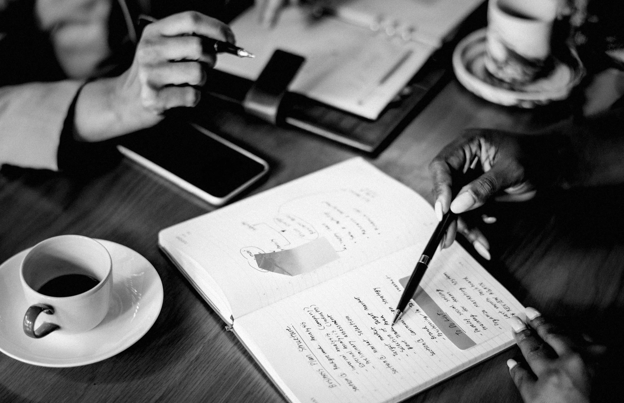 Black and white photo of two people sitting at a table, one writing in a notebook and the other holding a pen, with a cup of coffee, a phone, and a few papers on the table.