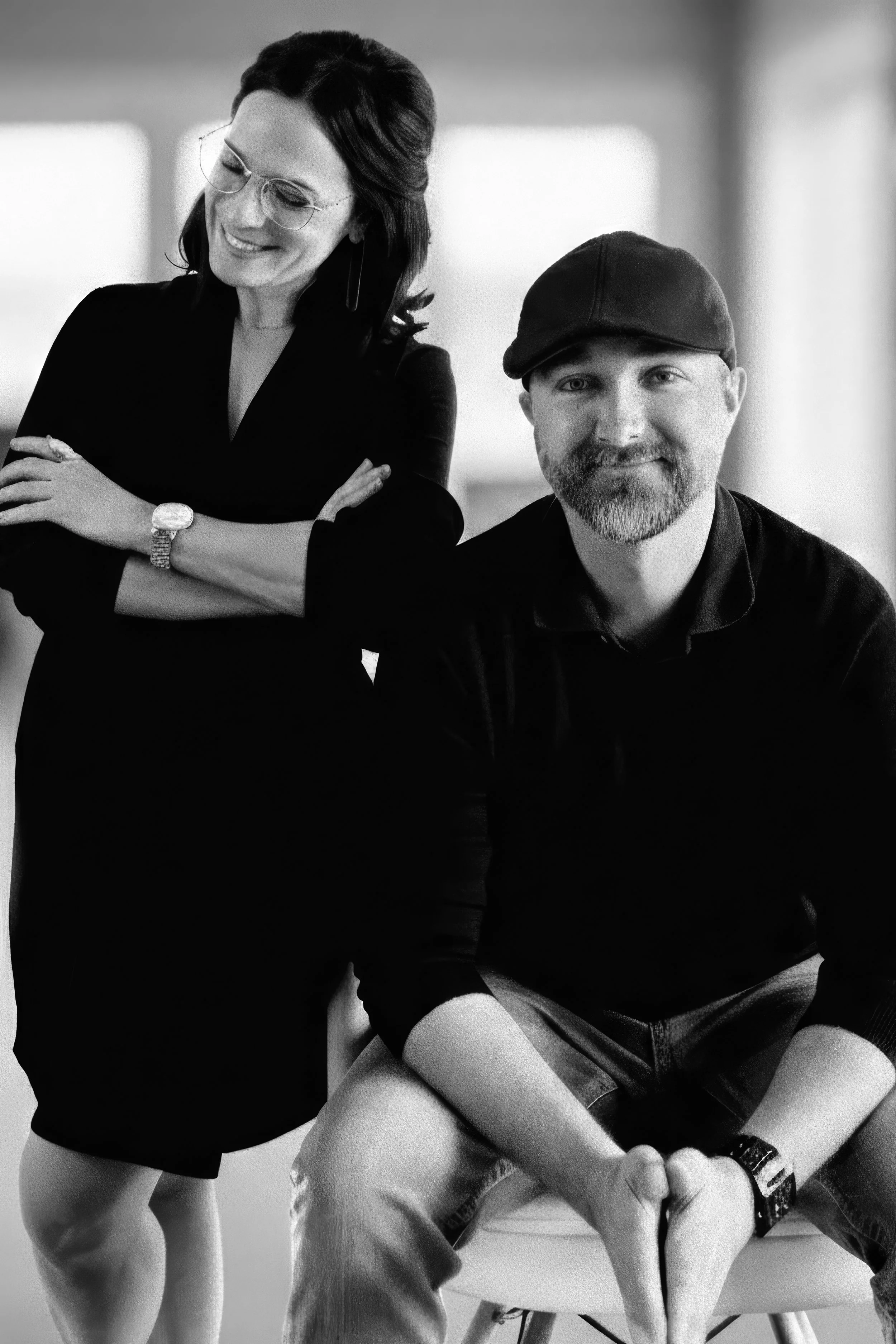 A black and white photo of a woman with dark hair and glasses standing next to a seated man with a beard, wearing a cap, both smiling.