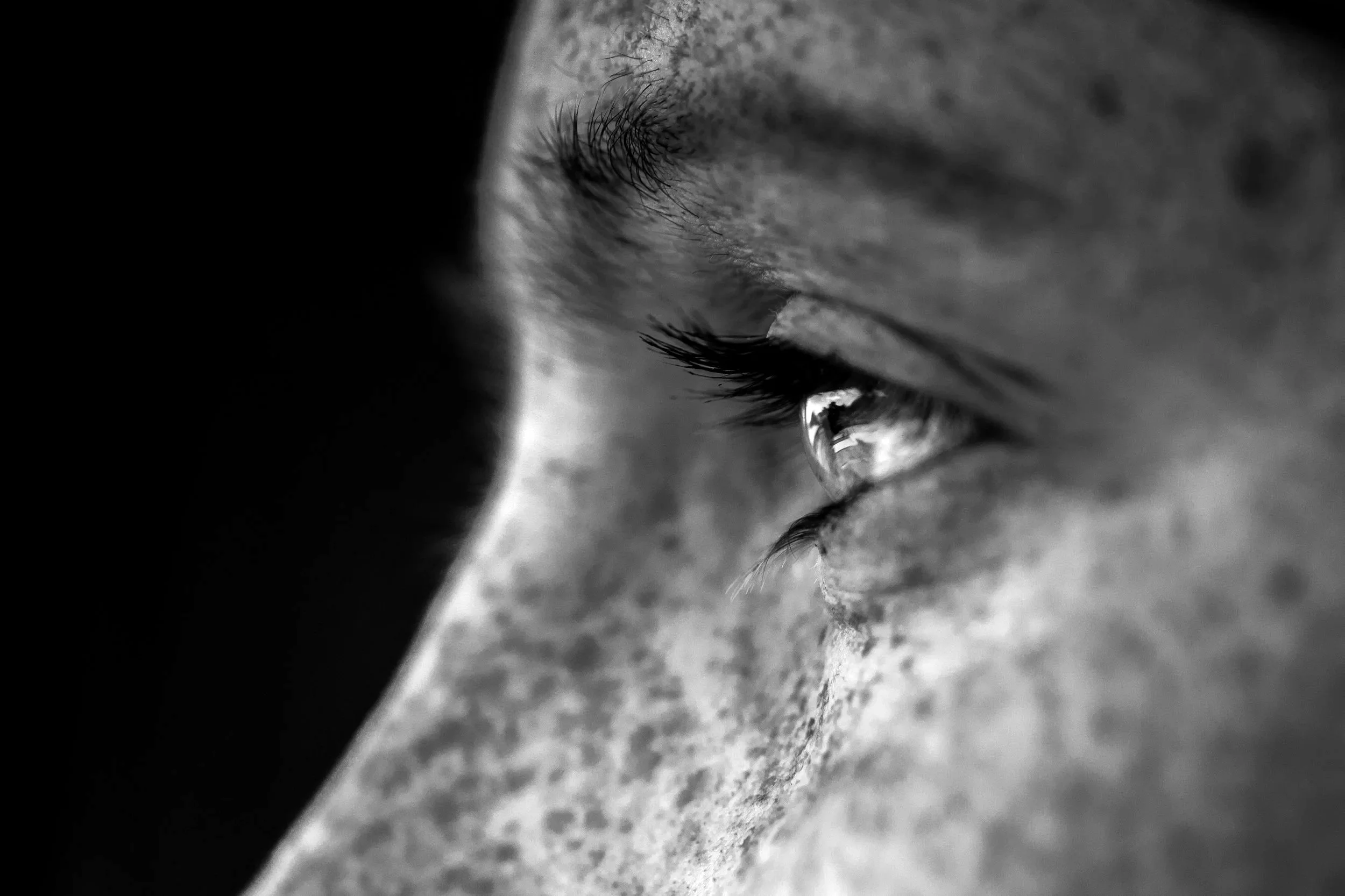 Close-up black and white photograph of a person's face focusing on the eye, with visible eyelashes and a tear falling from the eye.