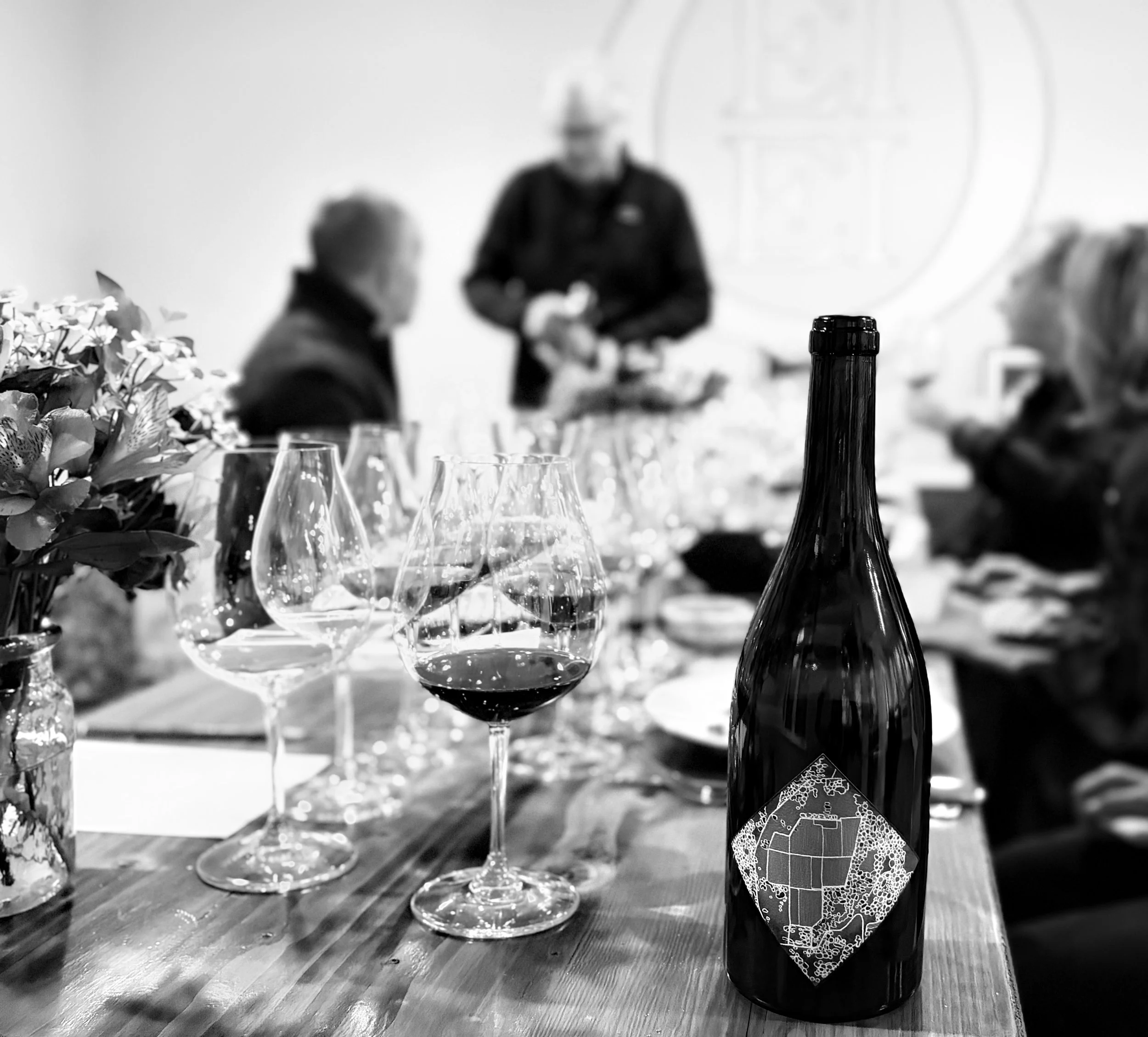 A black and white photo of a dinner table set with wine glasses, a wine bottle with a decorative label, and a vase with flowers. People are seated around the table, with one person standing in the background, possibly serving or preparing food.