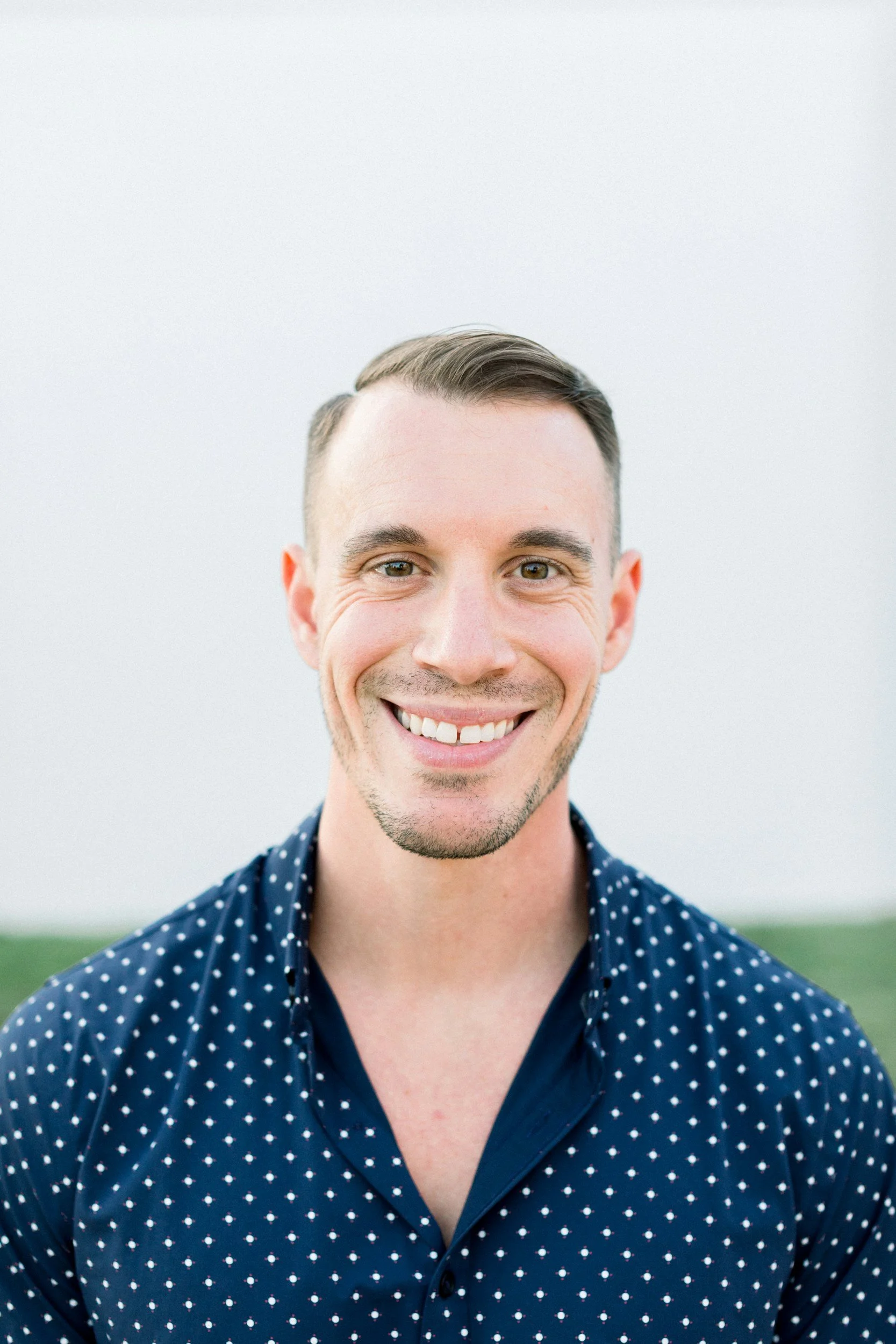 Close-up portrait of a smiling man with short brown hair, light skin, wearing a navy blue shirt with white polka dots, outdoors with a plain background.