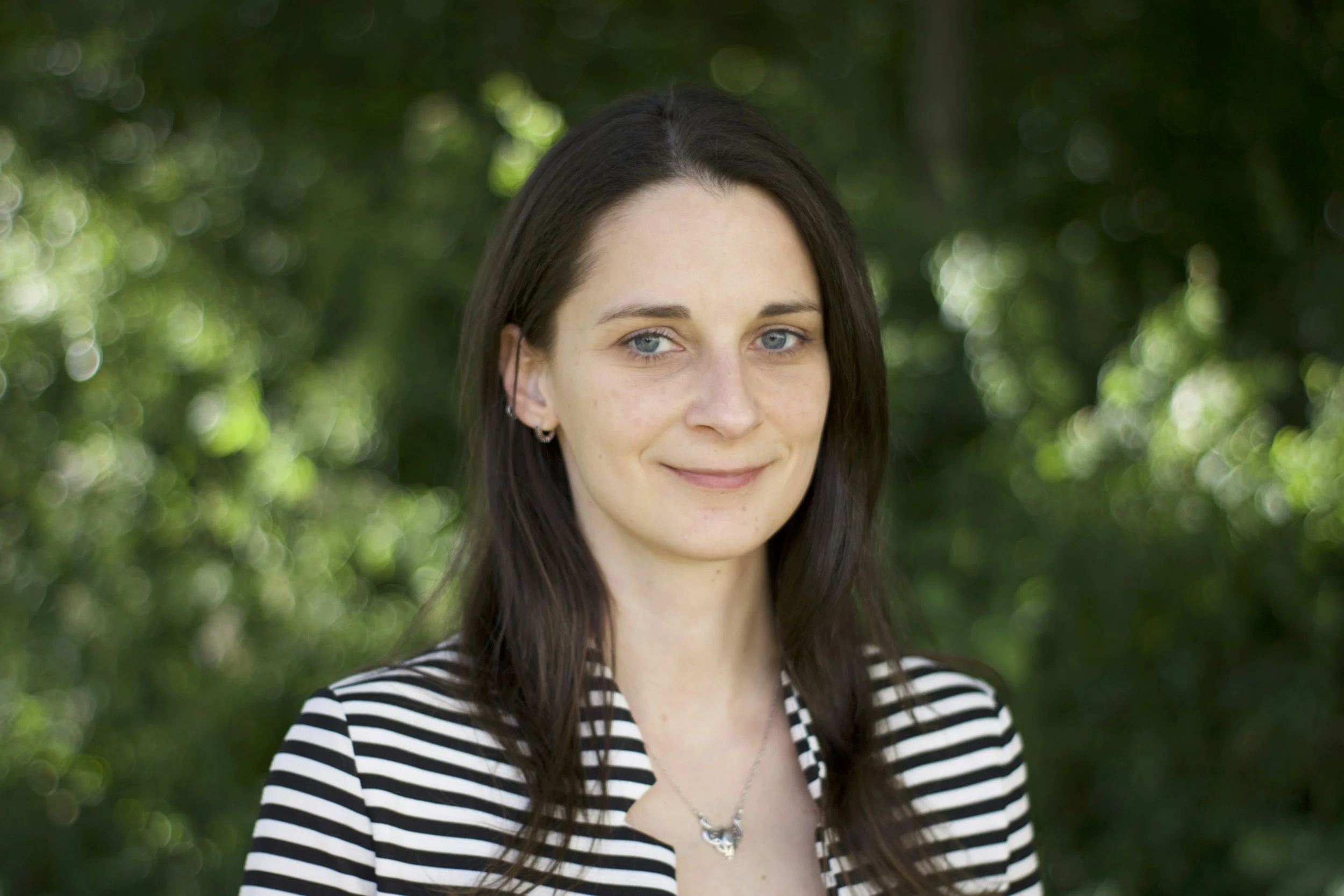 A young woman with long dark hair, blue eyes, wearing a striped black and white top, silver earrings and a necklace, standing outdoors with a blurred green foliage background.