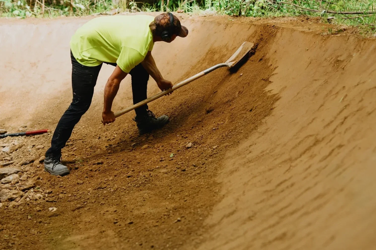 A person wearing a yellow shirt, black pants, and a cap is shoveling dirt in a trench. The individual is using a metal shovel and is working outdoors with green vegetation in the background.