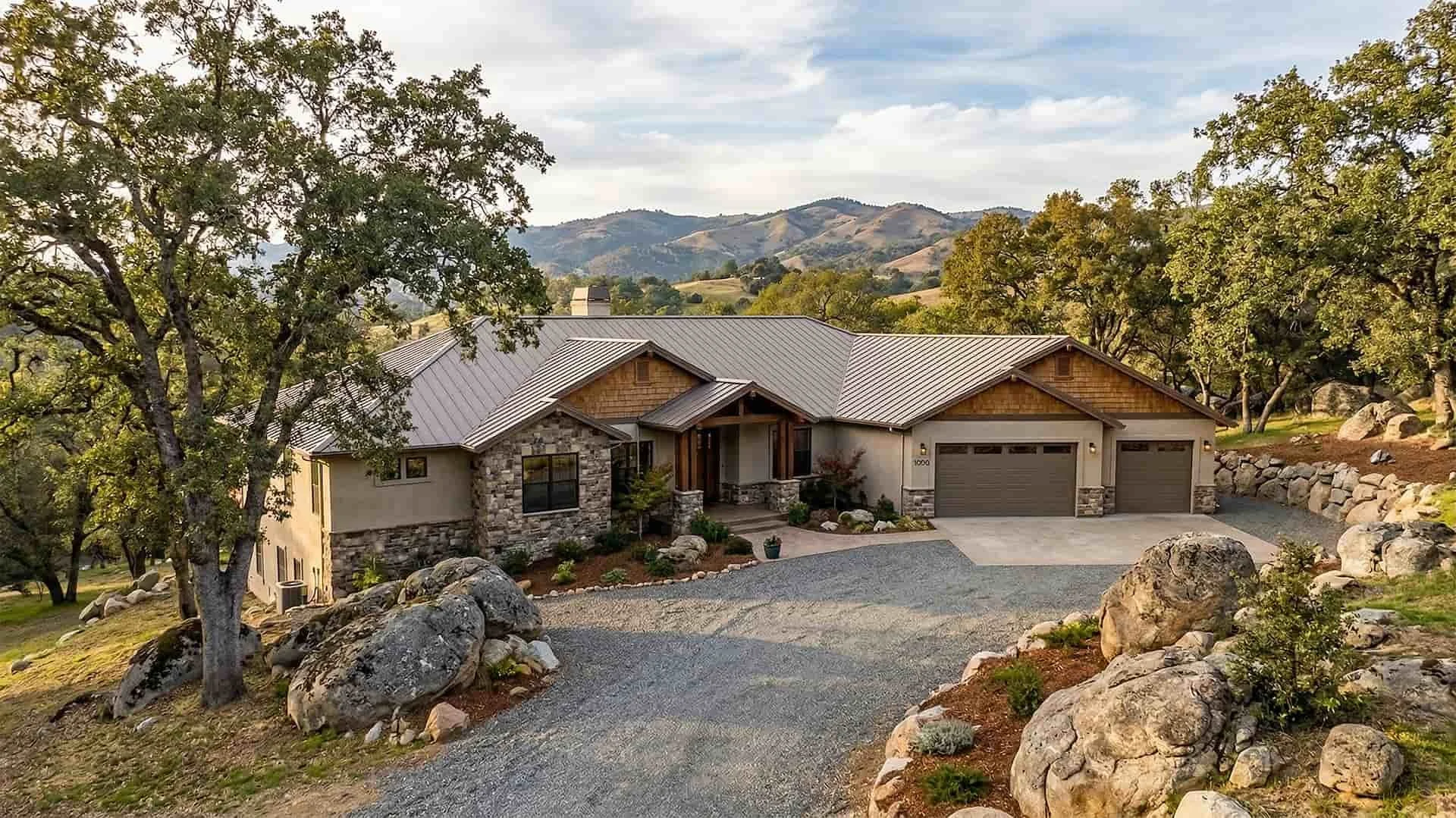 A modern house with a stone and stucco exterior, metal roof, and attached garage, surrounded by trees and rocks in a hilly landscape.