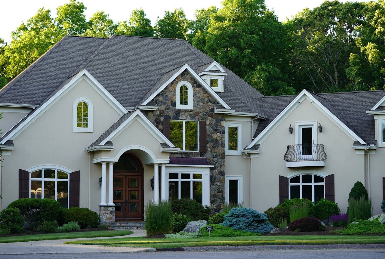 Front view of a large, elegant house with a well-maintained garden featuring various shrubs, trees, and rocks; the house has white walls, dark shutters, multiple gables, and stone and wooden accents.