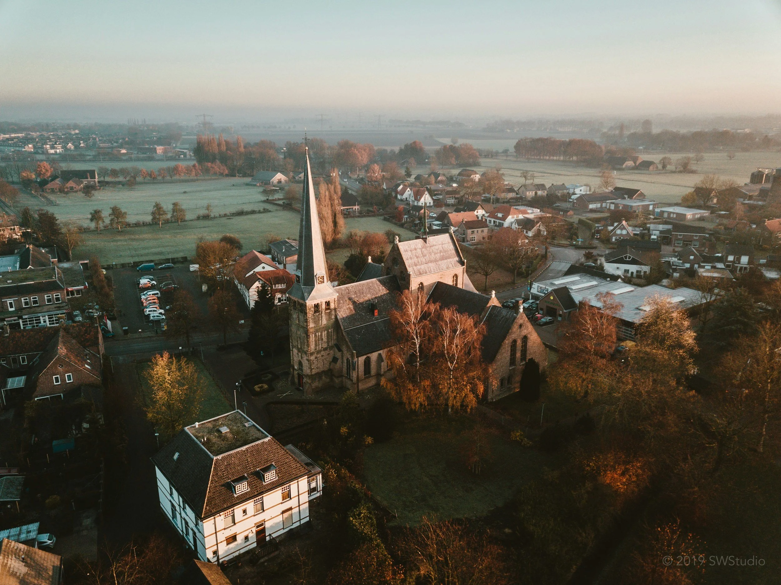 An aerial view of a small town with a church, residential houses with red and dark roofs, green fields, trees, and a river in the background under a clear sky.