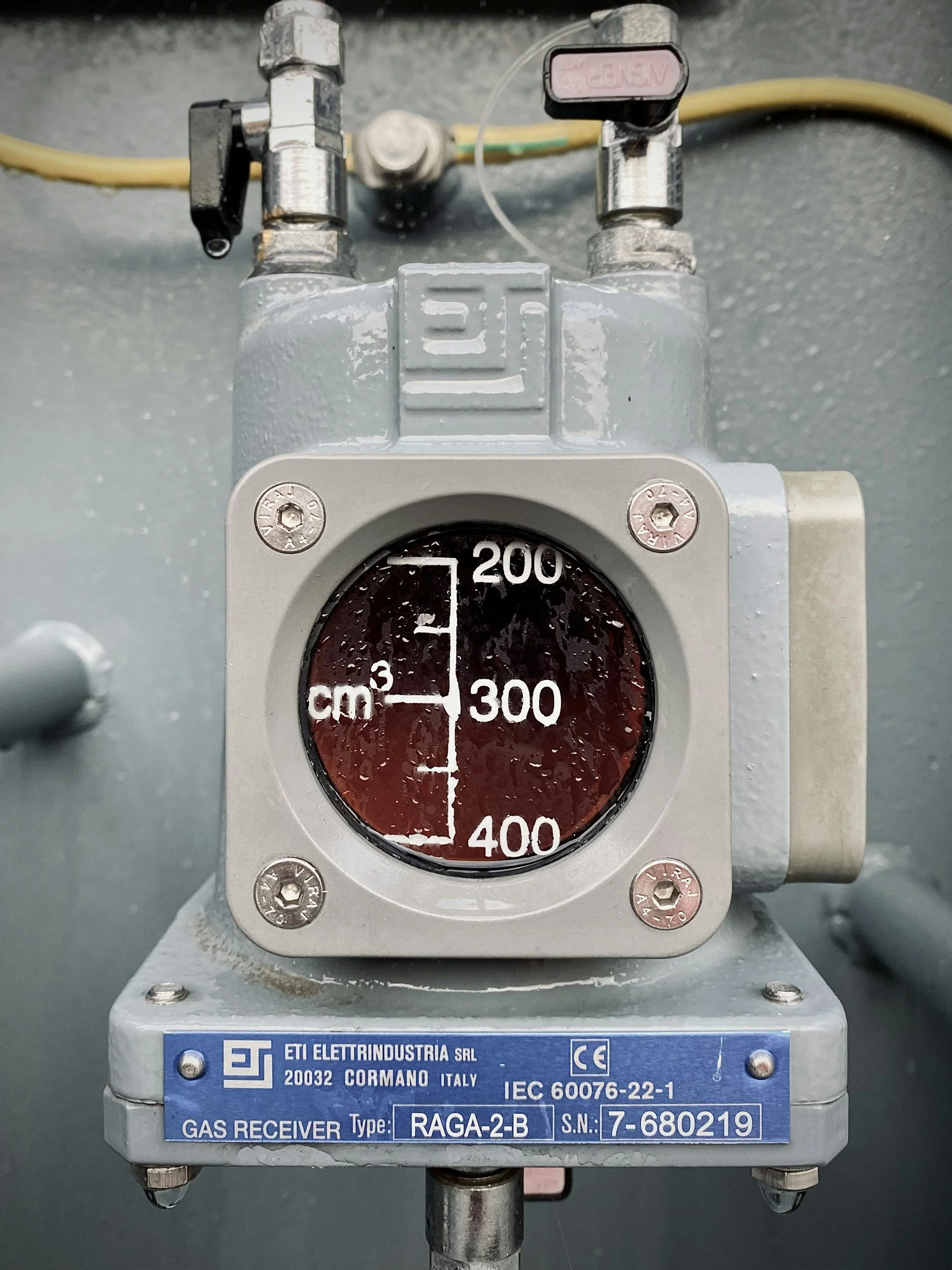 A gas receiver device manufactured by ETI Elettrindustra, labeled RAGA-2-B, with serial number 7-680219, mounted on a metallic frame. The device has a circular gauge with pressure readings from 0 to 400, with a red liquid inside. There are various pipes and fittings connected to it.