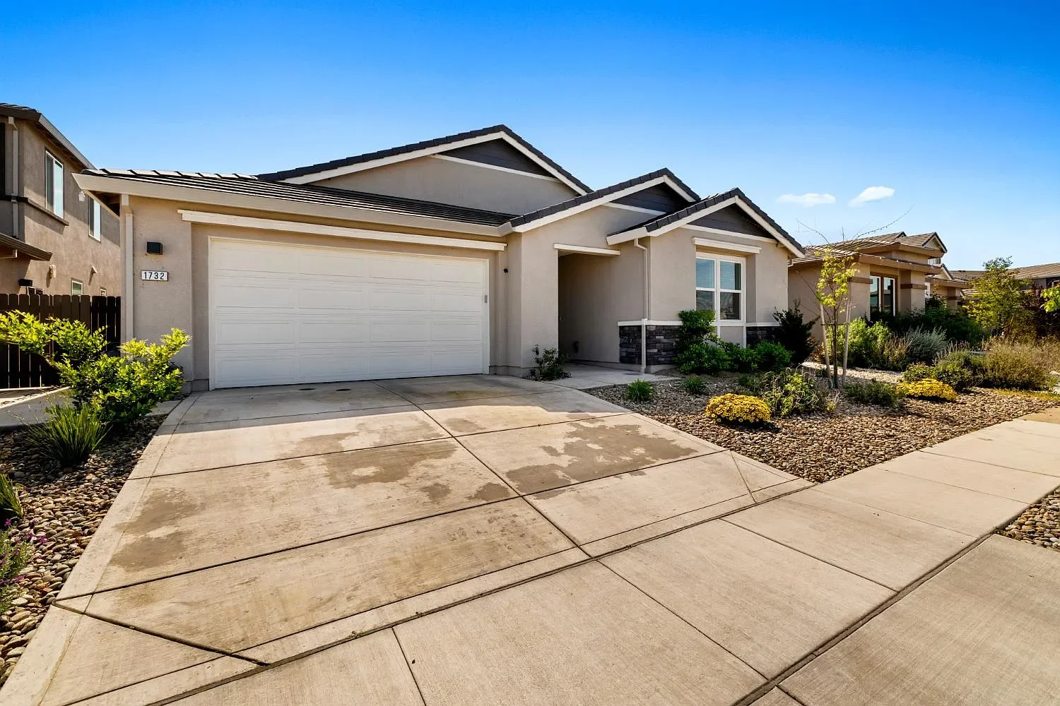 Modern single-story house with a concrete driveway, white garage door, and front yard landscaped with rocks, small plants, and trees under a blue sky.