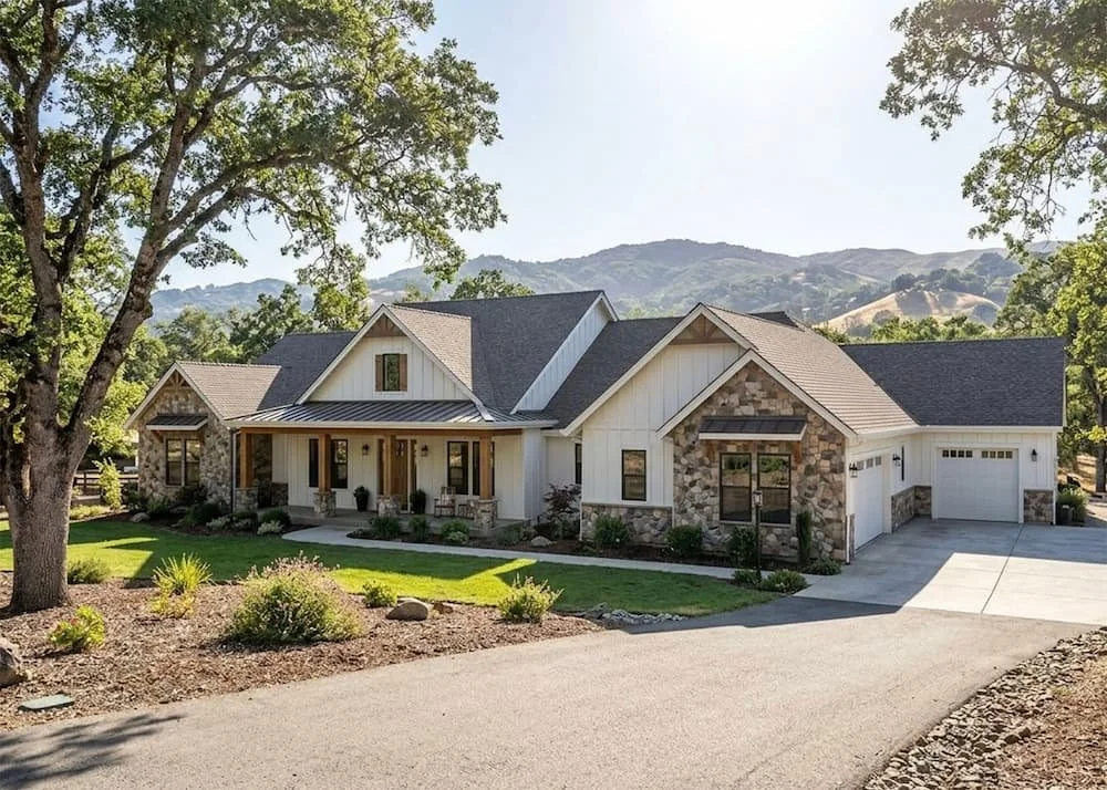 A modern single-family house with a combination of stone and white siding, surrounded by green trees and a landscaped yard, with a mountain range in the background under a clear sky.