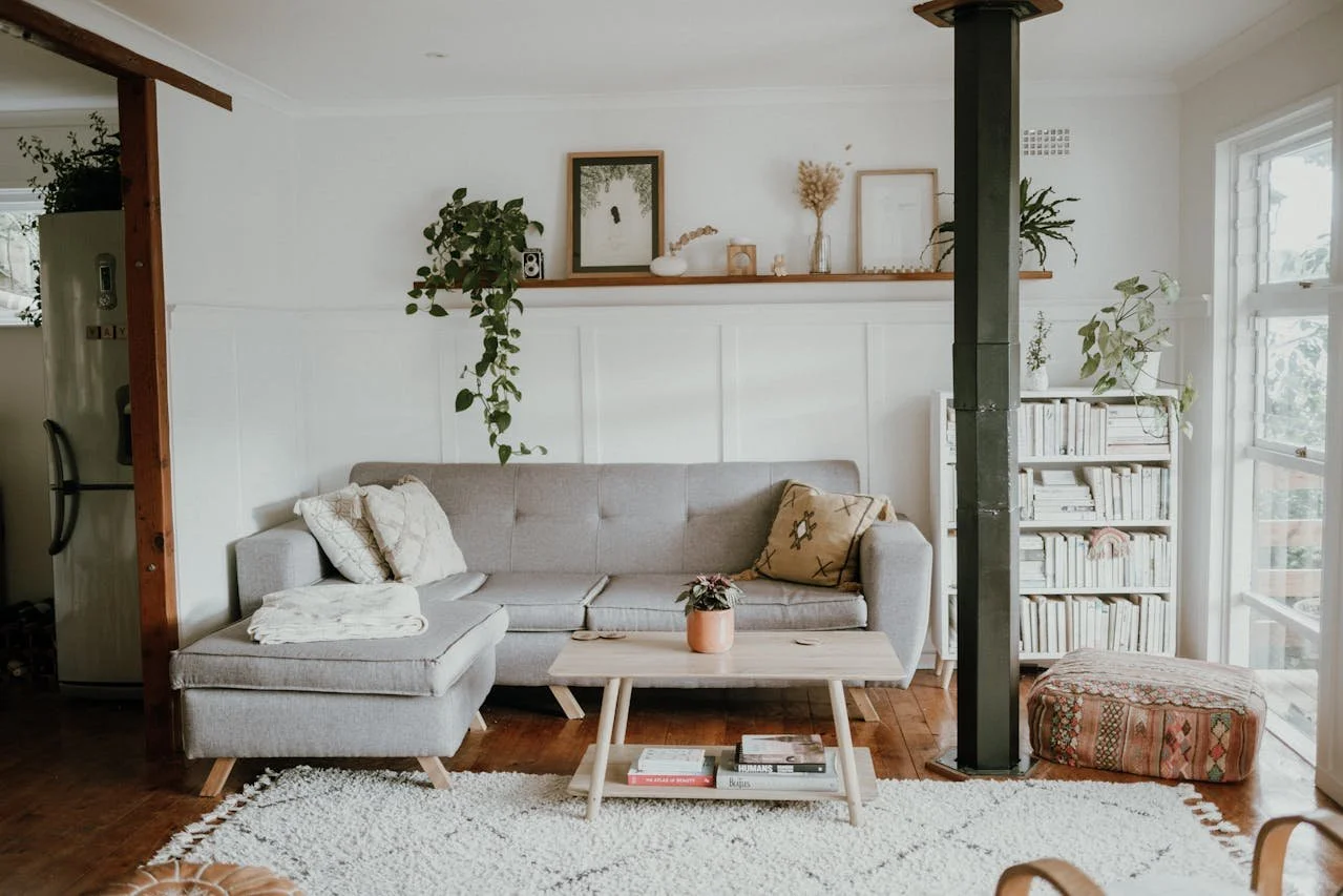 A cozy living room with a light gray sectional sofa adorned with decorative pillows, a small coffee table holding a plant, a white bookshelf filled with books, a window allowing natural light, and plants decorating the space.