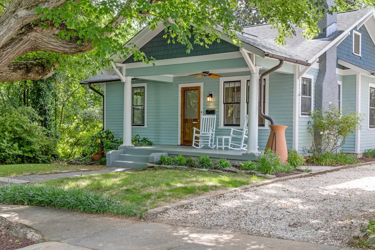 Front porch of a light blue house with a wooden door, two white rocking chairs, and a small table, surrounded by greenery and trees.