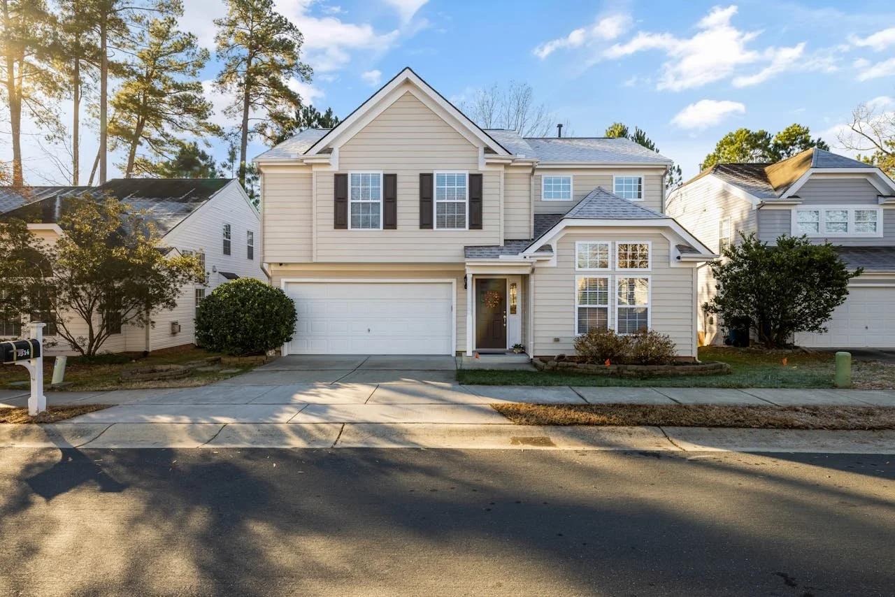 Two-story suburban house with cream-colored siding, black shutters, and a front lawn, with neighboring houses on either side, sunset light, and blue sky with a few clouds.