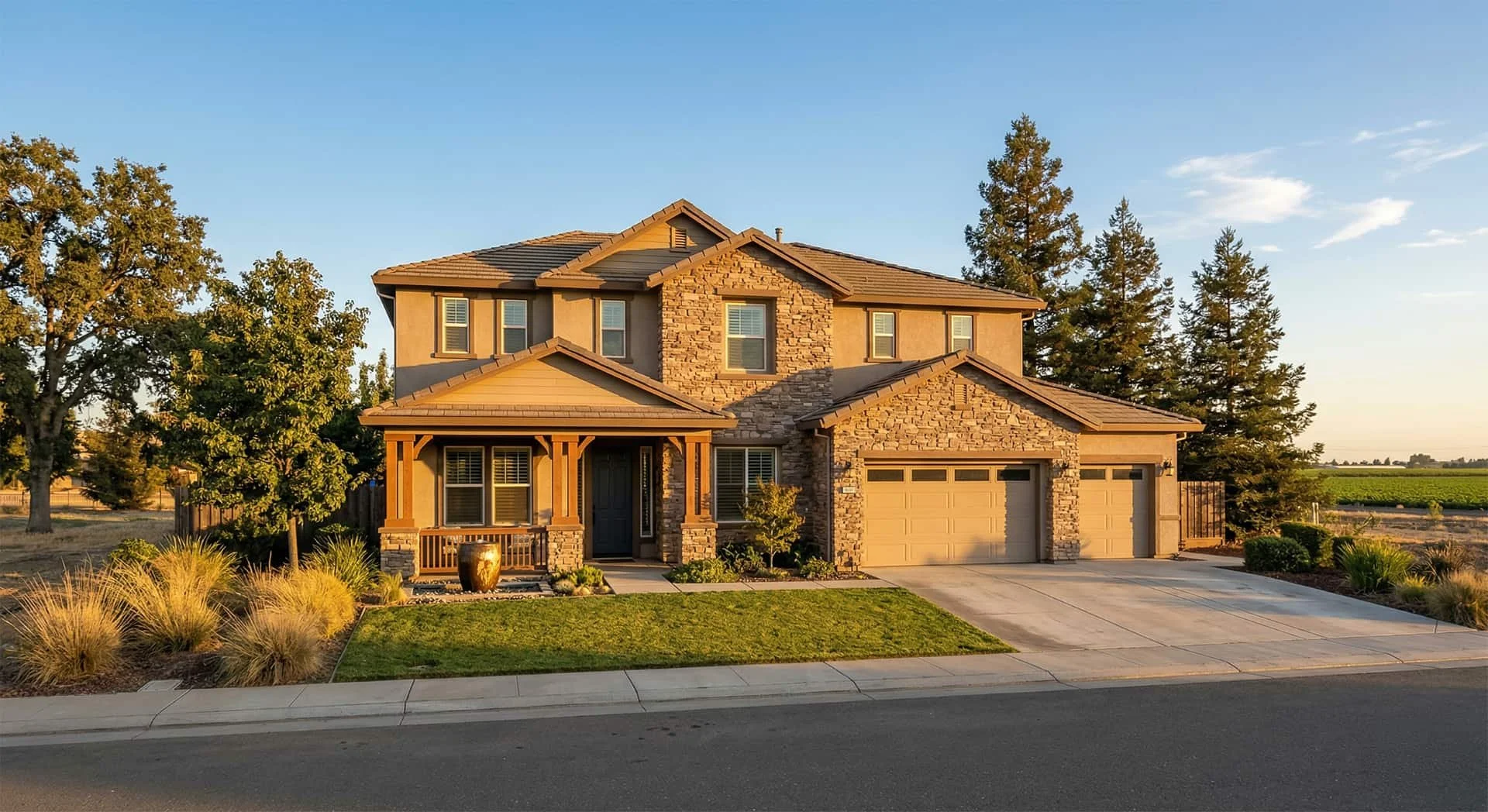 A modern two-story house with a stone and stucco exterior, a tiled roof, a covered front porch, and an attached two-car garage, surrounded by trees and landscaped yard during sunset.