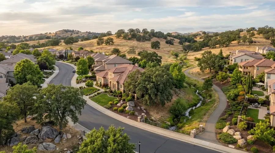 A residential neighborhood with houses, green lawns, trees, and a small creek with walking paths, set in a hilly, semi-arid landscape.