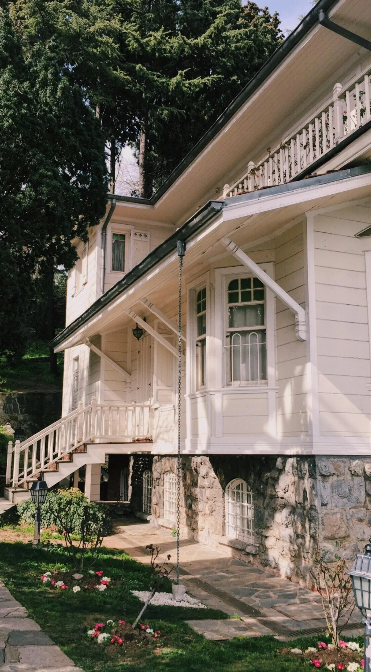 A two-story white house with a stone foundation, surrounded by a garden with pink and white flowers. The house has a staircase leading to the front door, a small balcony with a white railing, and multiple windows. Tall trees are visible in the background.