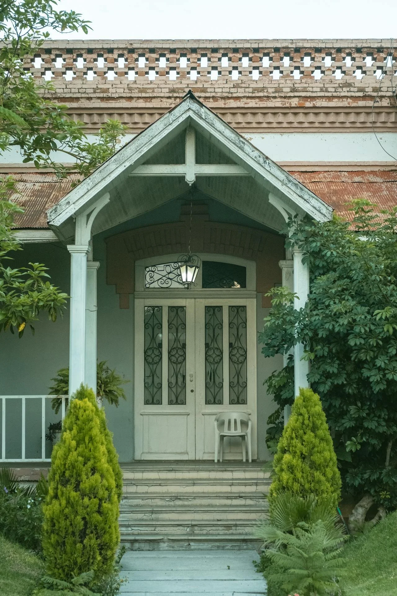 Front porch of a house with a white door and ornate ironwork, surrounded by green plants and trees, with stairs leading up to the entrance.