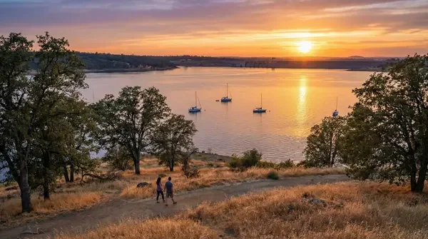 A scenic view of a lake at sunset with five sailboats anchored, surrounded by trees and grassy hills, with two people walking along a trail in the foreground.
