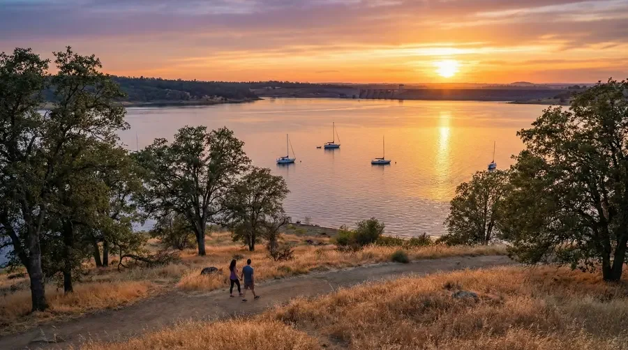 A scenic view of a calm lake at sunset with four sailboats floating on the water, surrounded by trees and dry grass, with two people walking along a dirt path in the foreground.