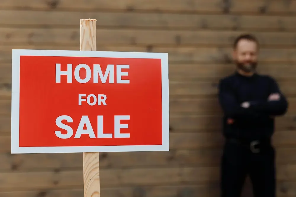 Sign that reads 'Home for Sale' in front of a man standing with arms crossed, blurred background of a wooden wall.