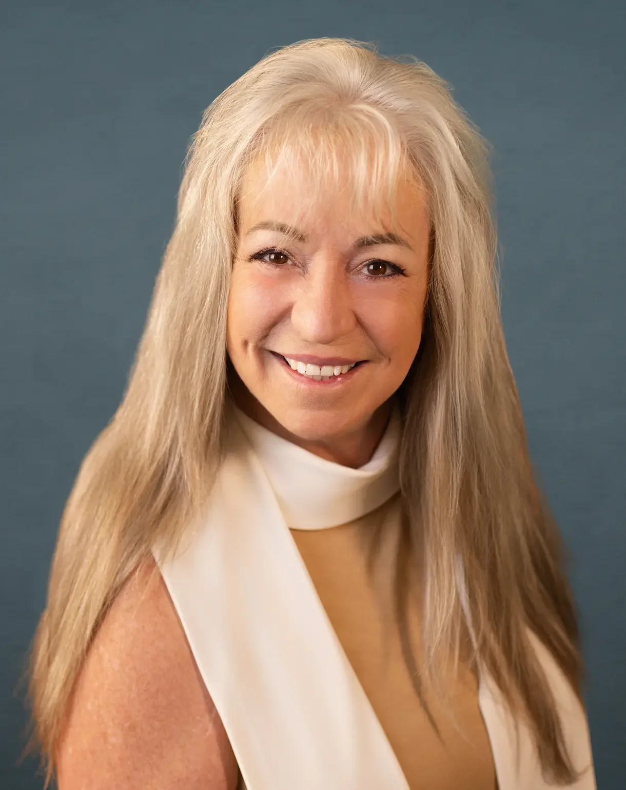 A smiling middle-aged woman with long blonde hair, wearing a cream-colored sleeveless top, poses against a solid blue background.