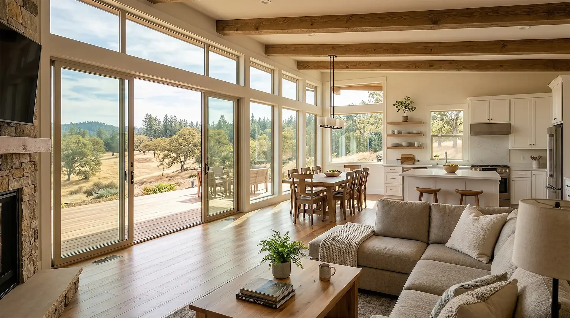 Open-concept living and dining area with large floor-to-ceiling windows showcasing a scenic outdoor landscape of trees and a hill. The space features a beige sectional sofa, wooden coffee table, dining table with six chairs, white kitchen cabinets, and a small potted plant. A sliding glass door opens to a patio with outdoor seating, overlooking the natural scenery.