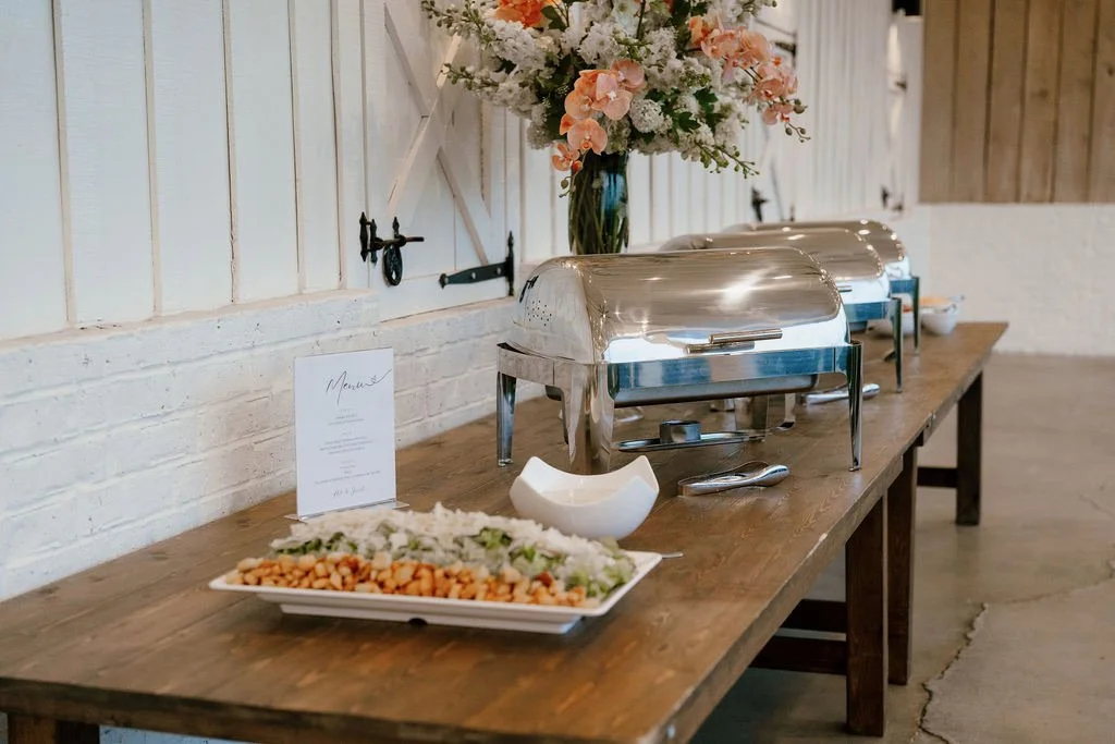 Buffet table with chafing dishes, a vase with pink and white flowers, a menu card, and a tray of salad with chickpeas.
