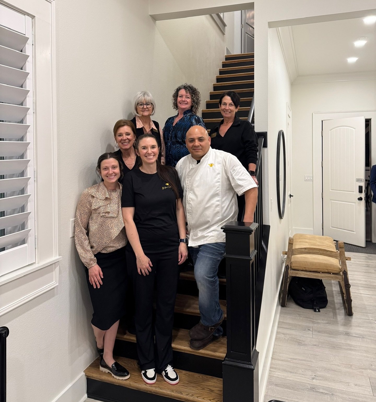 Group of seven people gathered on a staircase in a home, posing for a photo. The group includes six women and one man, some in black uniforms and a chef with a white coat, all smiling.
