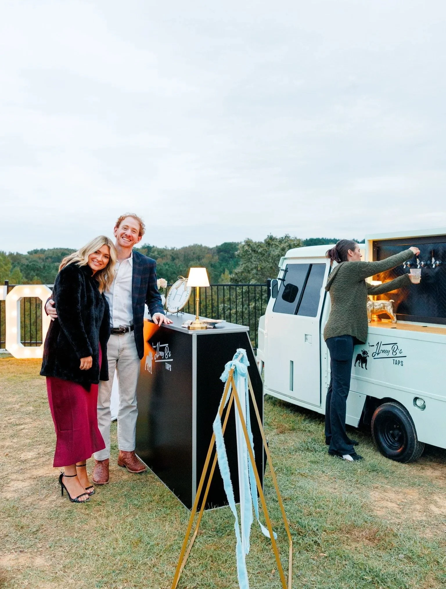 Two couples standing near a black table and a white food truck at an outdoor event, with a woman serving drinks from the food truck.