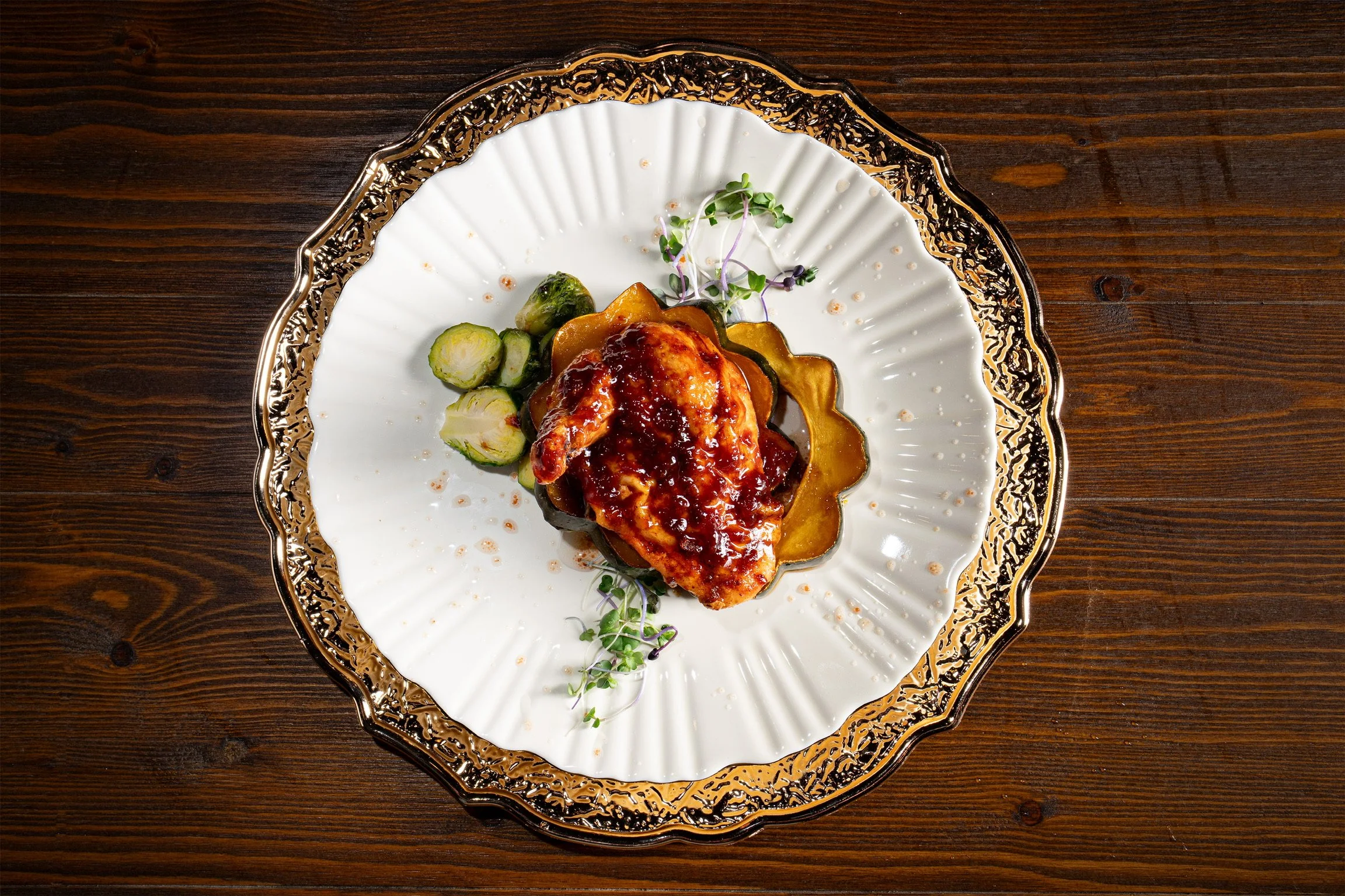A plated dish with roasted vegetables, including zucchini and a sauce-covered main item, garnished with microgreens, on a decorative white and gold plate on a wooden table.