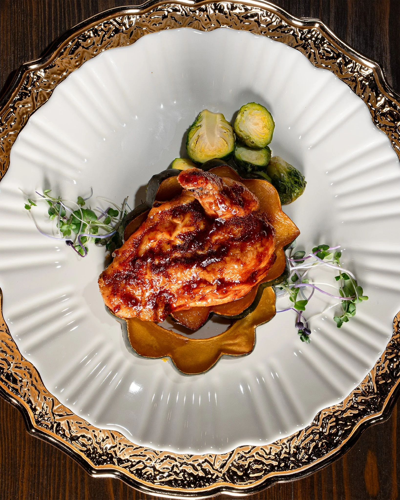 A plated dish with a piece of grilled chicken in tomato sauce, served with cooked Brussels sprouts and microgreens, on an ornate silver-rimmed white plate.