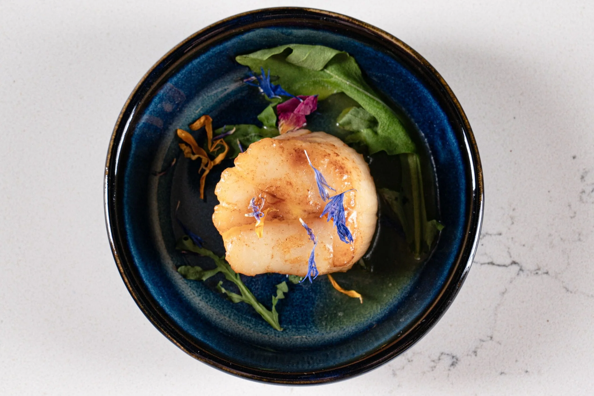 Seared scallop served on a bed of greens with edible flowers in a dark blue bowl.