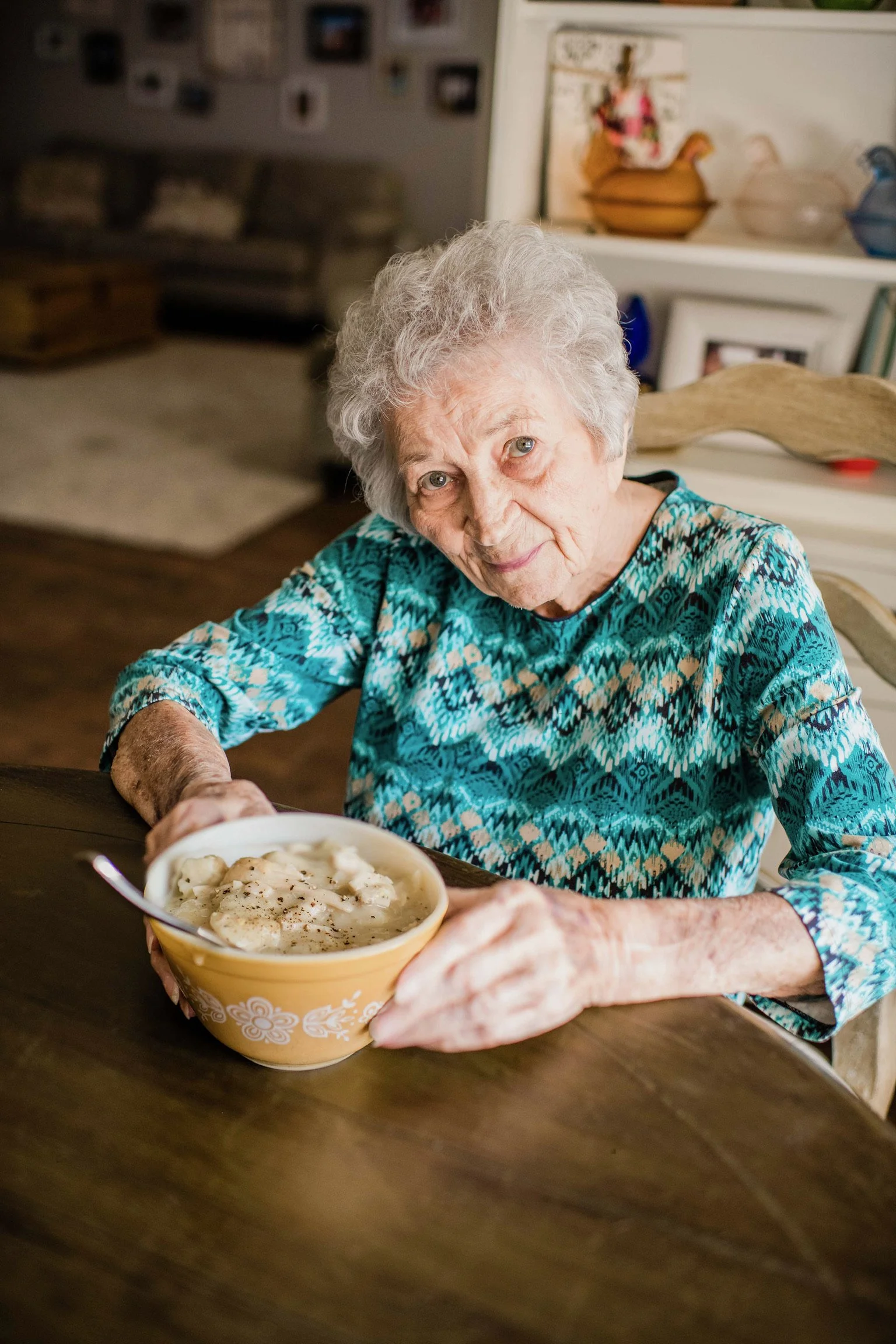 An elderly woman with curly gray hair and blue eyes sitting at a wooden table, holding a bowl of banana pudding and looking at the camera.