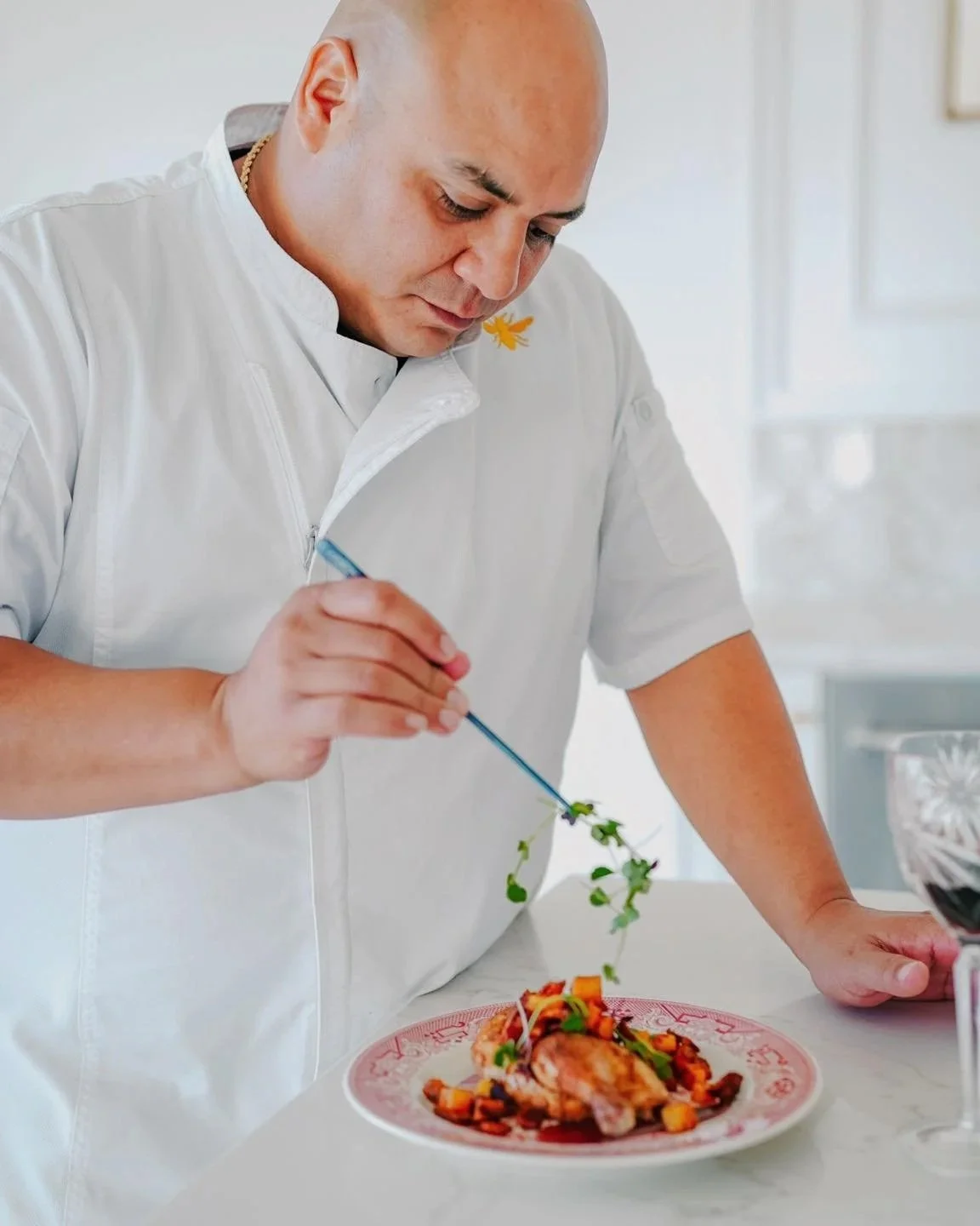 Chef garnishing a dish with herbs in a kitchen setting, with a glass of red wine nearby.