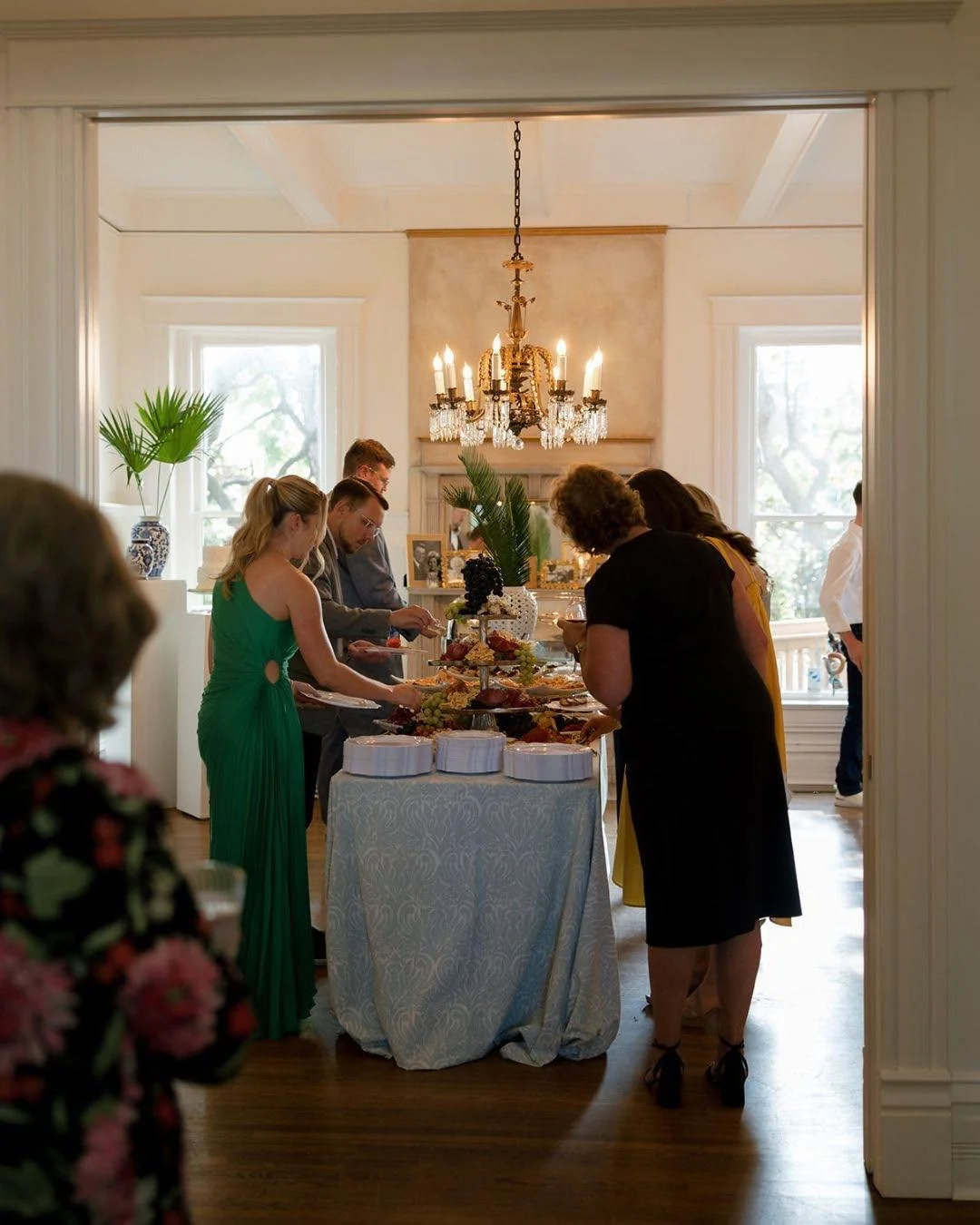 People gathered around a table with food, at a social event in a well-lit room with large windows and a chandelier.
