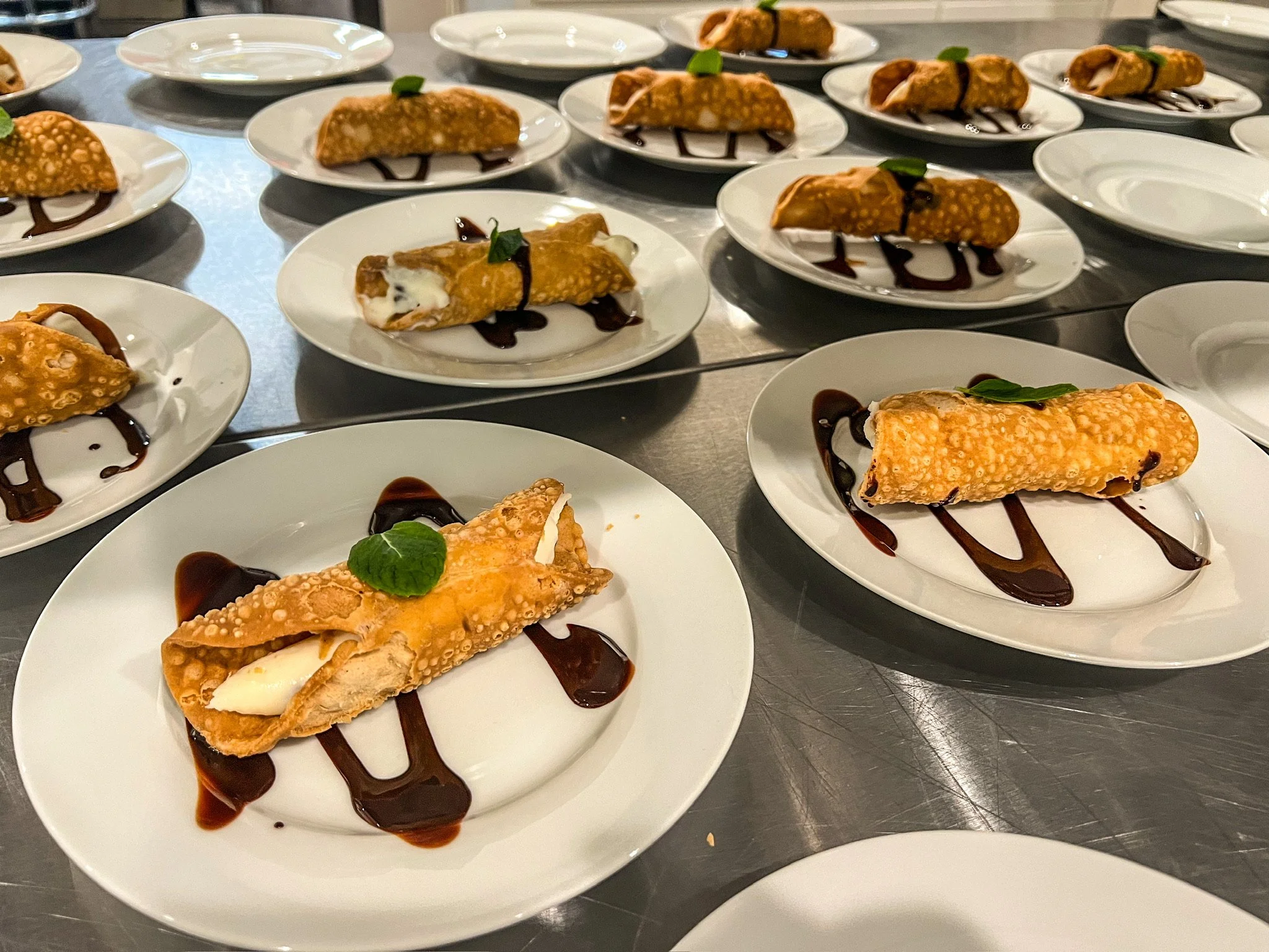 Several plates of fried dessert cannoli with chocolate sauce and mint garnish on a stainless steel table.