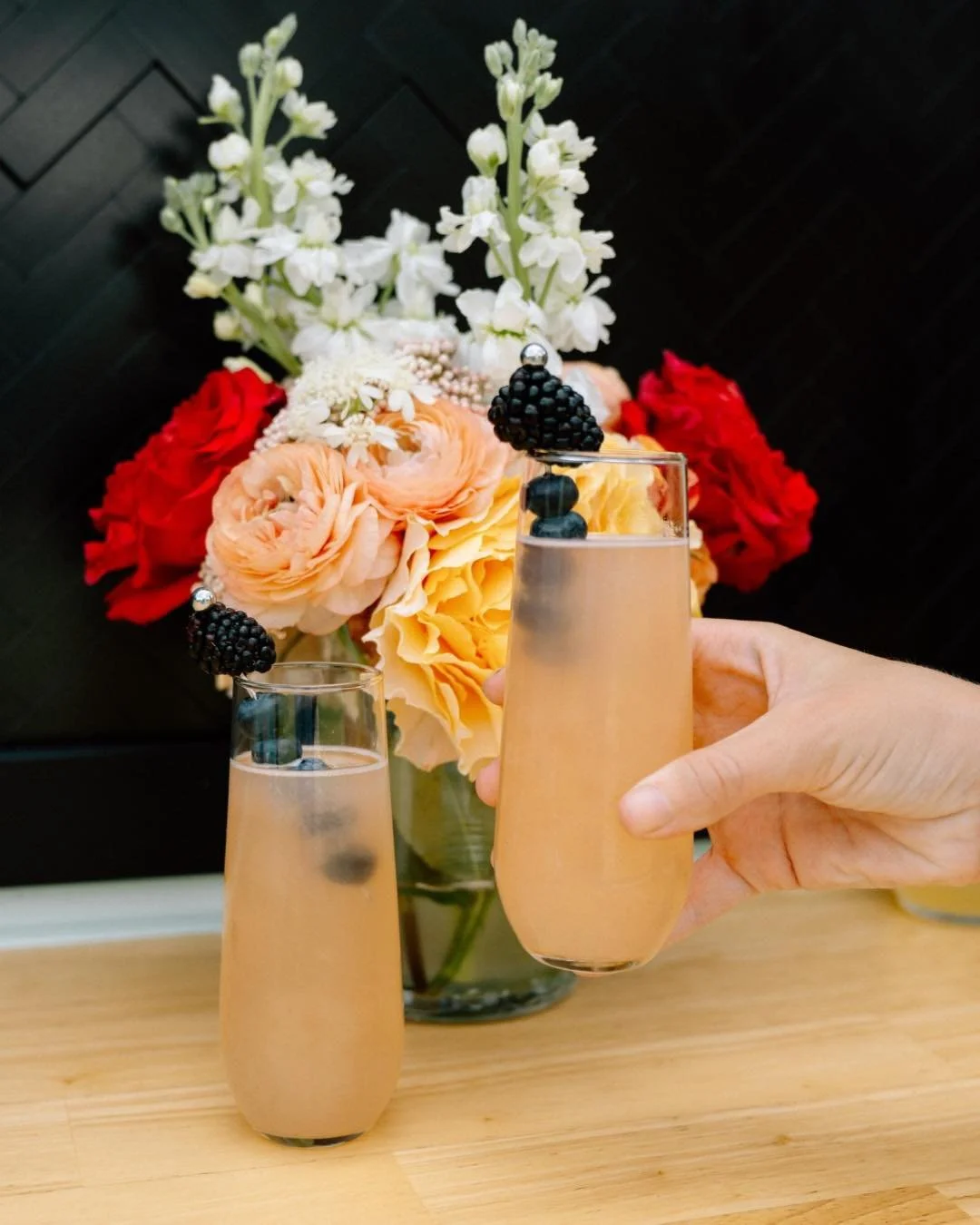 Two glasses of pink champagne with blackberries as garnish, held by a hand in front of a vase of red, peach, and white flowers on a wooden surface.
