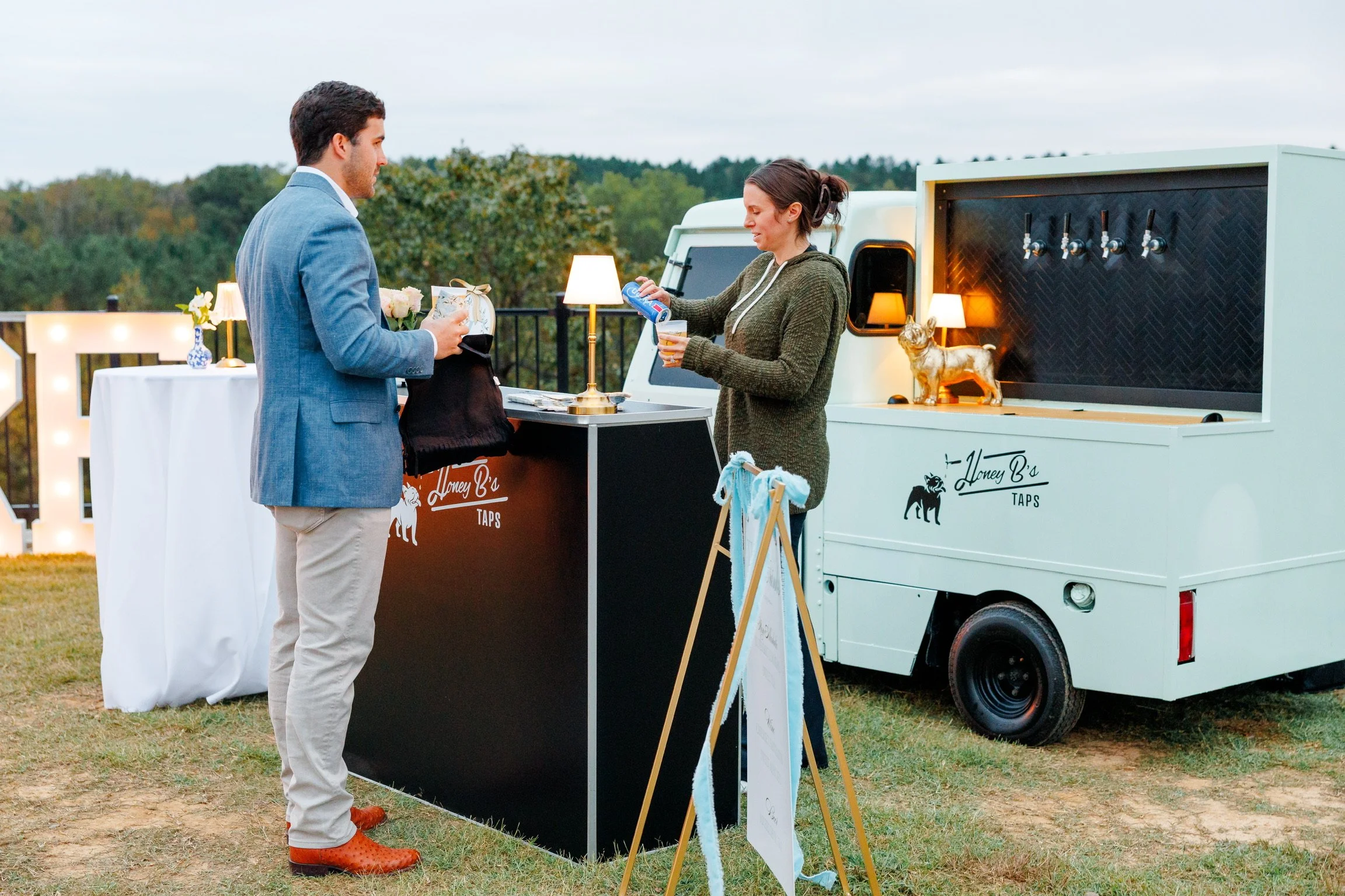 A man in a blue blazer and beige pants standing at a black and white drink stand, interacting with a woman in a green hoodie behind the stand at a mobile tap bar labeled Honey B's Taps, outdoors during evening with trees in the background.