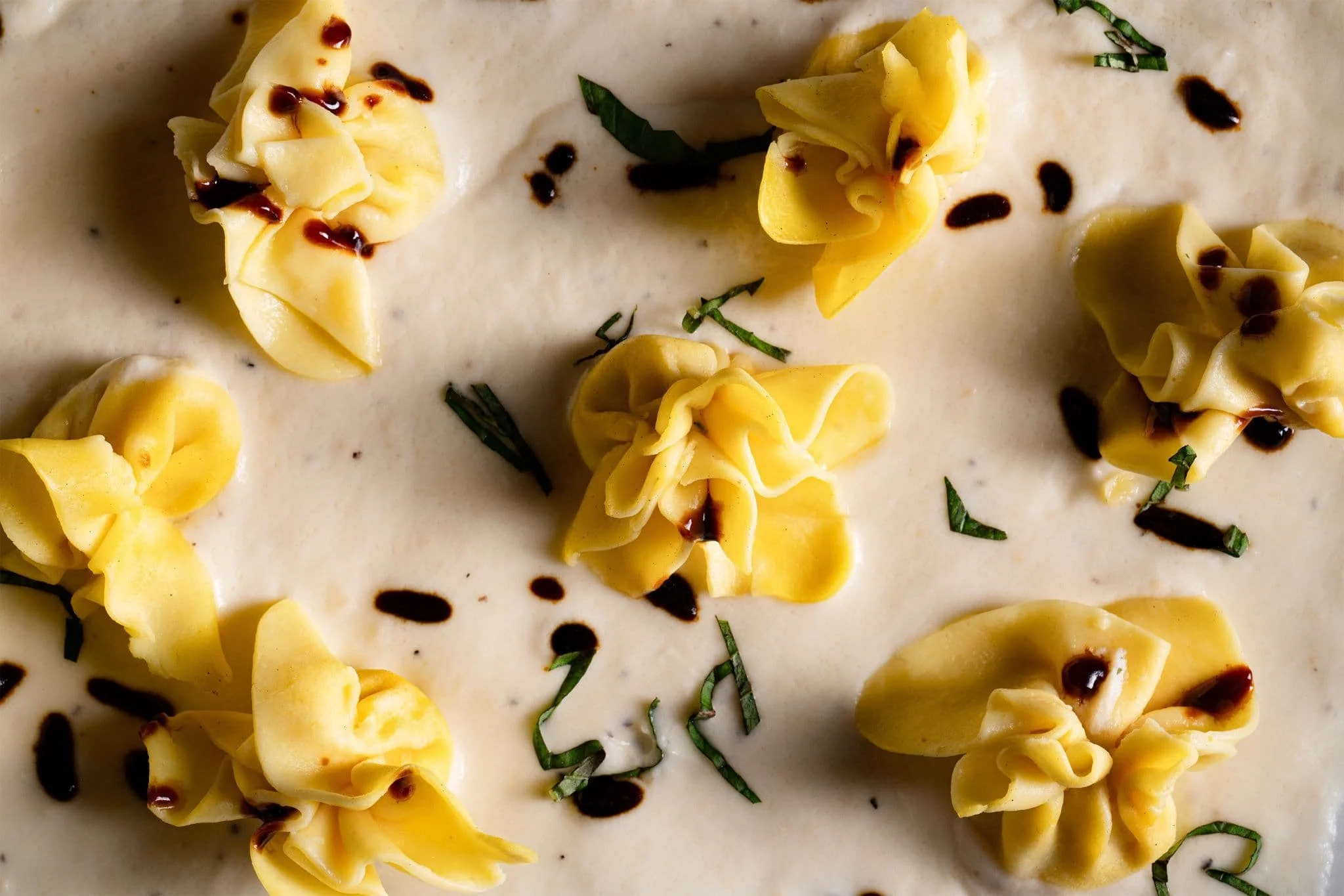 Close-up of tortellini pasta in a creamy white sauce, garnished with chopped herbs and drizzled with dark balsamic or soy sauce.